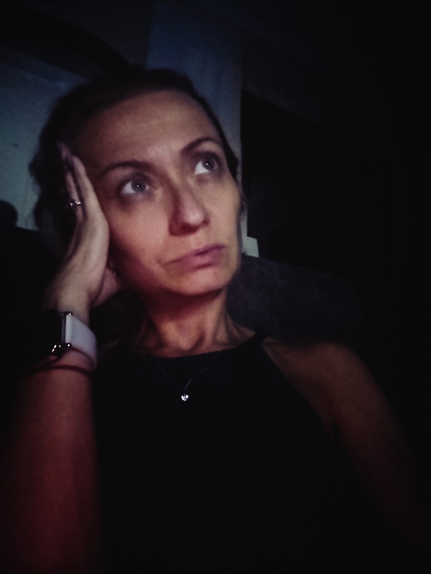 A woman is pictured in a dimly lit room, leaning her head on her hand and looking upwards. She is wearing a black top and a silver smartwatch. A small, delicate necklace with a pendant is visible around her neck. The background is dark and out of focus, with a hint of a textured wall. There are no identifying features in the image that would suggest a specific location in Moscow, Russia. The scene is still and quiet.