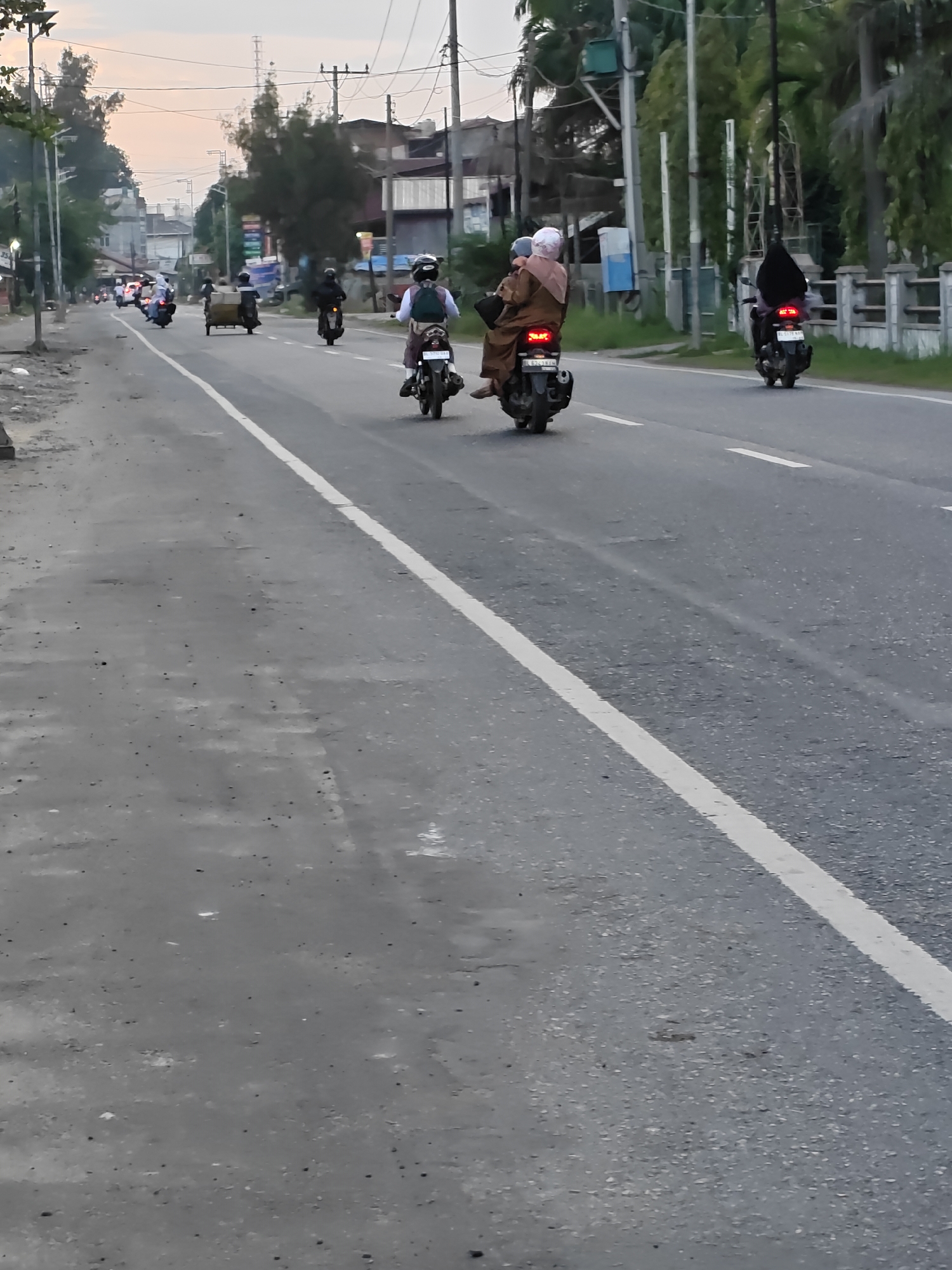 In Lhokseumawe, Indonesia, the image captures a street scene during what appears to be the late afternoon or early evening, as indicated by the soft, warm light. The road, a mix of asphalt and gravel, stretches into the distance, bordered by a white line on the right side. Along the left side of the road, a rougher, dirtier shoulder is visible, with some debris and patchy vegetation.
The background reveals a typical urban or semi-urban environment with various buildings and structures. Trees, some with dense foliage, line the sides of the road, and utility poles with wires are prominent. The buildings are varied in style and condition, suggesting a developed but possibly older area.
Several motorcycles are present, indicating common mode of transportation. In the foreground on the right, a motorcycle is partially visible, with its rear wheel and part of the exhaust system showing. Further down the road, two people are riding on a motorcycle, with the passenger wearing a headscarf. Another motorcycle with a rider and a passenger in a headscarf is further ahead. On the left side of the road, a motorcycle with a rider wearing a helmet and backpack is visible, and behind it, another motorcycle is seen. Two more motorcycles are further down the road, one with a rider in a black headscarf. In the middle ground, a motorcycle with a sidecar is traveling in the same direction as the other vehicles. In the far distance, more motorcycles are visible, suggesting a moderate amount of traffic. The overall atmosphere is one of everyday life and movement in a bustling Indonesian town.
