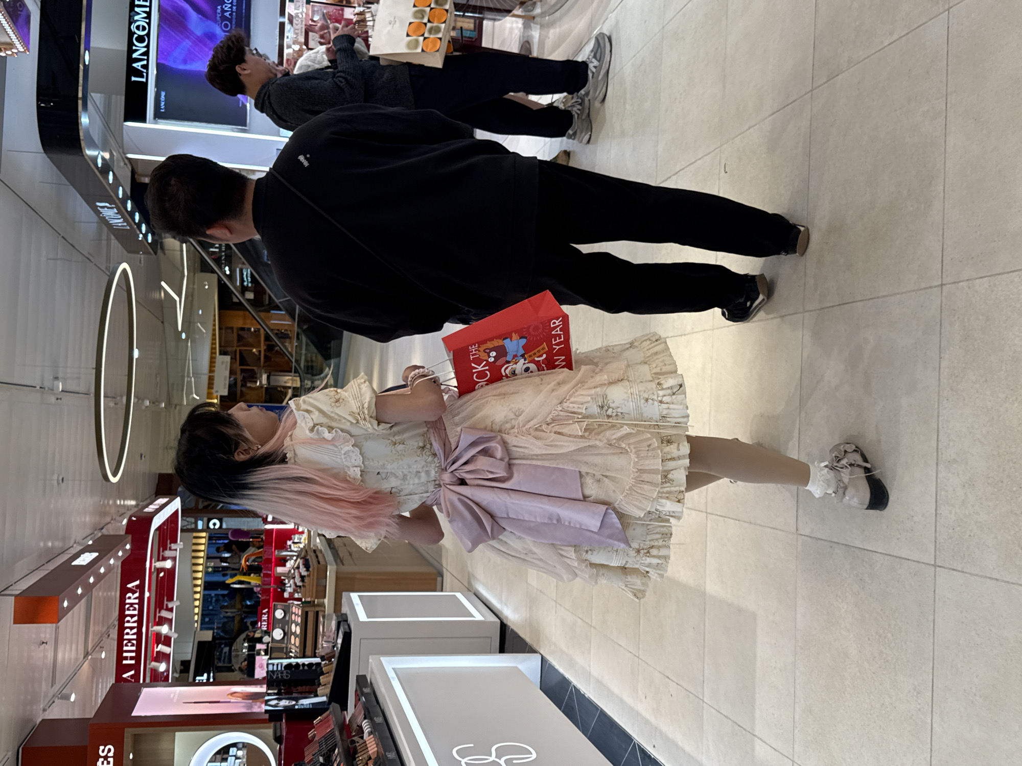 A photo of an individual in a dress with a ribbon and a red shopping bag standing in what appears to be a shopping mall. Two other individuals are visible further in the distance. Signage for "LANCOME" and "A HERRERA" is visible in the background. Cosmetic products are also displayed on shelves near where the individual is standing.