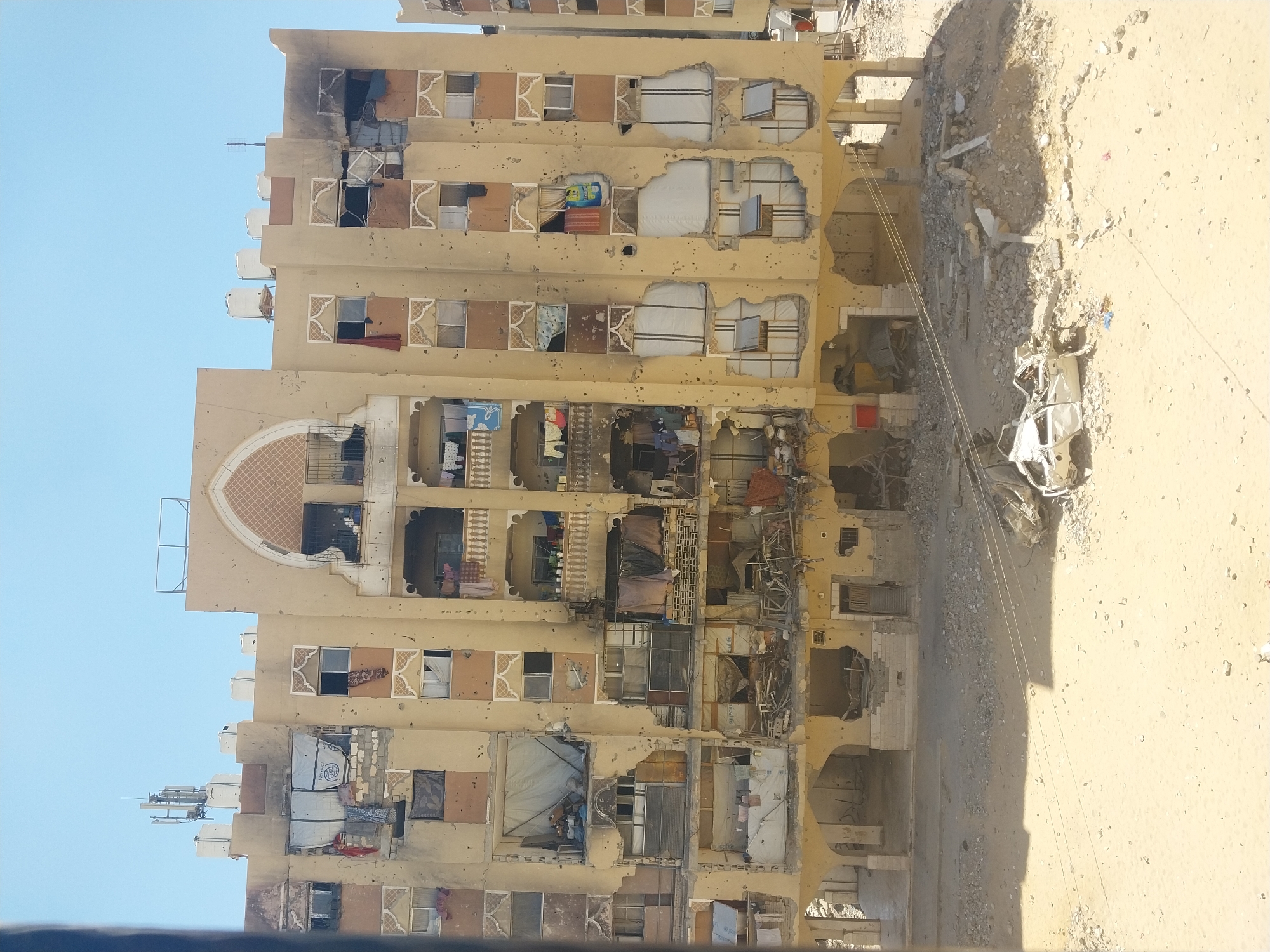 The image depicts a severely damaged apartment building in Khan Yunis, Palestinian Territories. The building, appearing to be several stories high, has sustained extensive damage, with numerous holes and craters visible on its facade, likely from shelling or explosions. Several windows are broken, and some apartments appear to have collapsed or been gutted. Debris is scattered across the ground in front of the building, including rubble, broken concrete, and mangled metal structures. A heavily damaged white car lies on its side amidst the debris, indicating a recent destructive event. Clothes are hanging from some of the balconies, suggesting that people may still be inhabiting the structure or have recently lived there. The overall scene conveys widespread destruction and devastation.