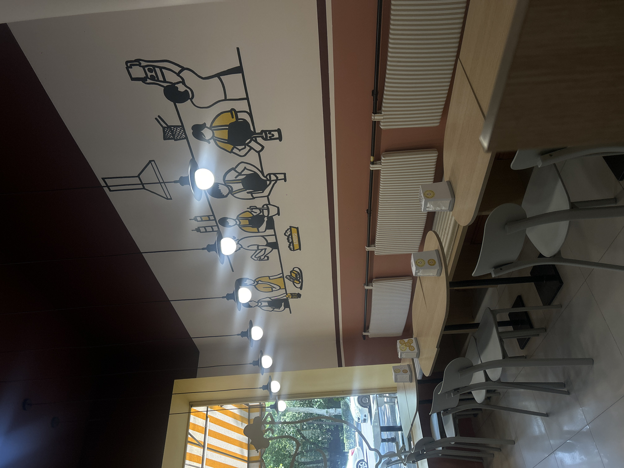 This is an indoor shot of a restaurant with a black ceiling and white walls. The walls are decorated with a mural of cartoon figures preparing food. There are several tables and chairs in the foreground, with striped banquettes along the wall. A window with yellow and white striped blinds is visible on the left side of the image, showing trees and cars outside. The overall ambiance is casual and modern. The establishment is located in Tbilisi, Georgia.