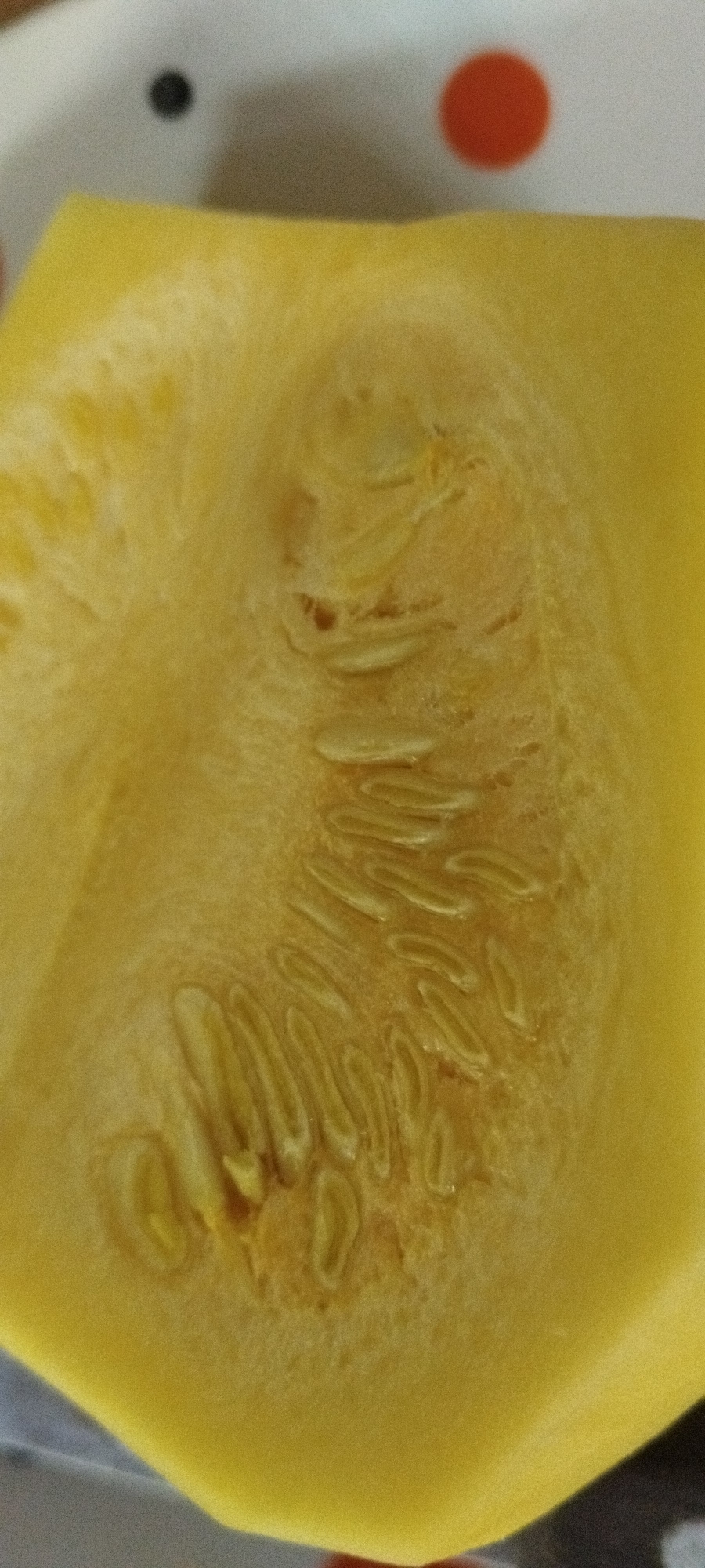 A close-up, macro shot focuses on the cut interior of a bright yellow squash. The flesh is a uniform yellow, with a slightly textured surface. In the center of the squash, a cluster of pale seeds is visible, embedded within a stringy, fibrous material. The seeds are elongated and appear smooth. The background is blurred, but suggests a white surface with a few scattered dark and red circular shapes, possibly part of a plate or placemat. The lighting is even, highlighting the texture and color of the squash. There is no indication of the location in Yaroslavskaya, Russia.