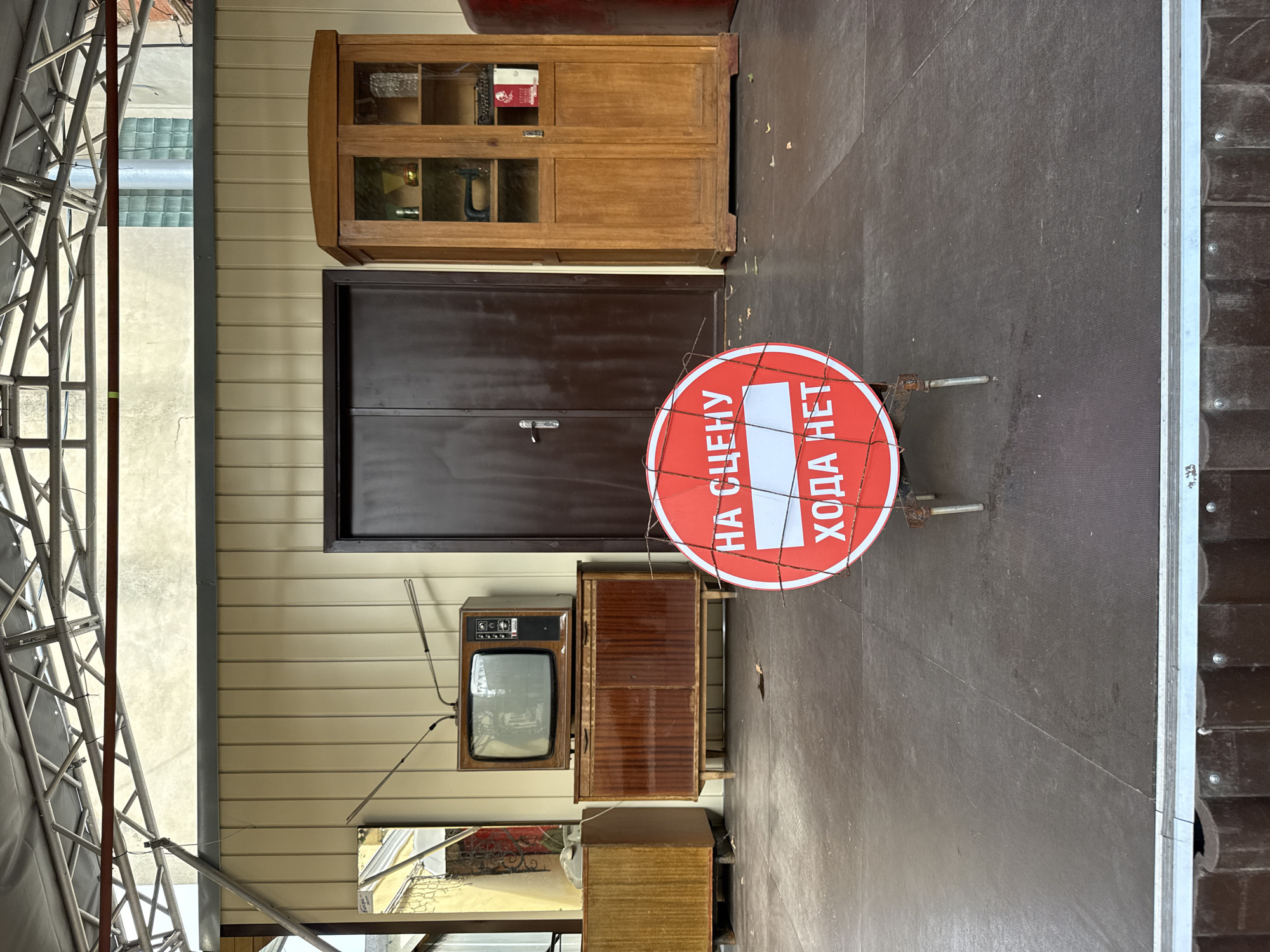 The image depicts an outdoor stage or platform in Nizhny Novgorod, Russia, set up with retro-style furniture and a prohibition sign. In the background, a light-colored, vertically paneled wall serves as a backdrop, with a dark brown double door in the center. To the left of the door, a vintage television set sits atop a dark wooden cabinet with two doors. Further left, a mirrored wardrobe or tall cabinet is partially visible. To the right of the double doors stands a tall, wooden cabinet with glass doors, containing various objects, including what appears to be a sewing machine and books. In the foreground, a circular red and white prohibition sign is prominently displayed, secured by wire. The sign reads "НА СЦЕНУ" (On Stage) with a white horizontal bar over "ХОДА НЕТ" (No Entry). The platform itself is dark gray and appears to be constructed of wood or a similar material. The scene is set under a metal framed canopy.
