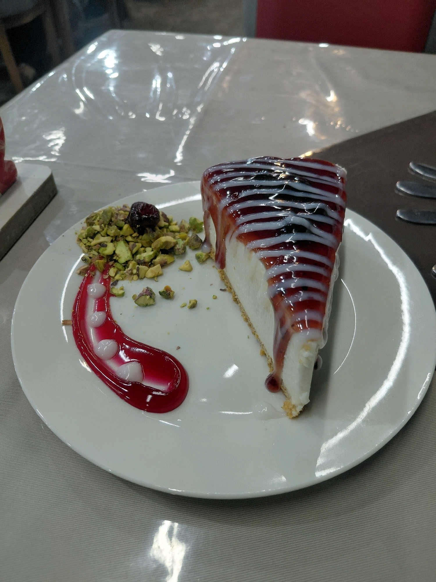 A slice of cheesecake is presented on a white plate. The cheesecake has a thick crust and is topped with a red sauce and white cream drizzled in stripes. To the left of the cheesecake, a pile of chopped pistachios is scattered, with a single dark red berry on top. A red sauce with white dollops is swirled on the plate beside the pistachios. The plate rests on a table covered with a light-colored cloth. In the background, a clear plastic tablecloth is draped over another surface, showing reflections of light. A blurry red chair back is visible behind the plastic. The scene appears to be indoors, possibly in a restaurant setting.