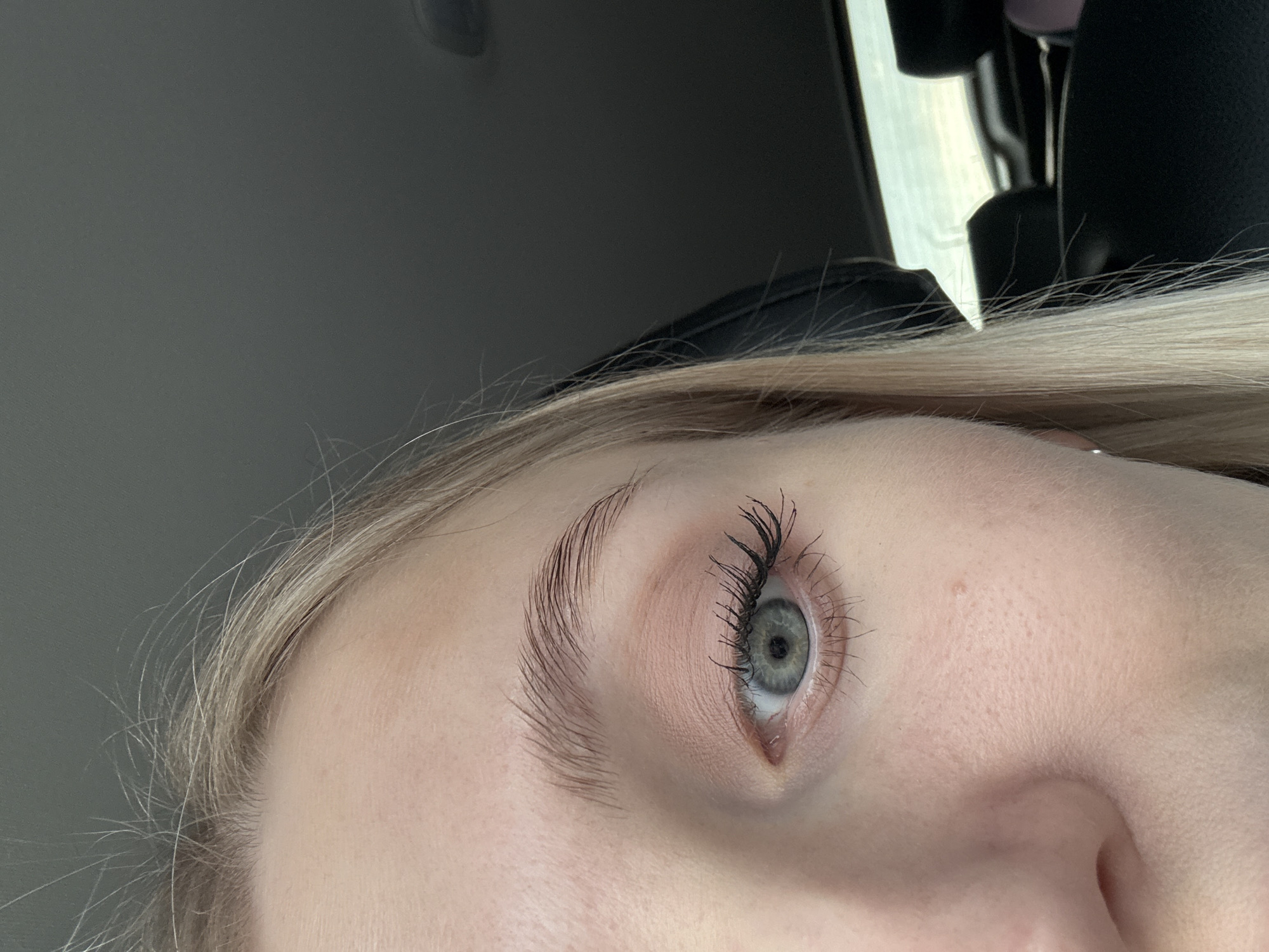 Close-up shot of a fair-skinned person's eye and brow, likely taken inside a vehicle. The eye is a light green-gray color and the person is wearing mascara. The eyebrow is light brown and well-groomed. Part of the nose is visible at the bottom of the frame, and blond hair is visible near the top of the frame. The background shows parts of the vehicle's interior: a dark gray ceiling, part of a headrest, and a glimpse of the outside through the car window. No information suggests this image was taken in Izhevsk, Russia.