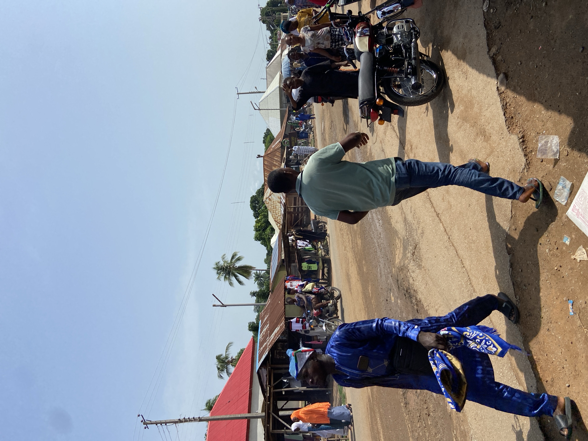 A street scene in Mararaba, Nigeria shows a sandy road with people and motorcycles. A man in a blue, patterned outfit walks toward the foreground, holding a folded blue cloth. Another man in a light green shirt and jeans walks in the opposite direction. Several people are on motorcycles further down the road, near small shops and buildings with metal roofs. Palm trees and power lines are visible in the background. The sky is light blue and mostly clear.