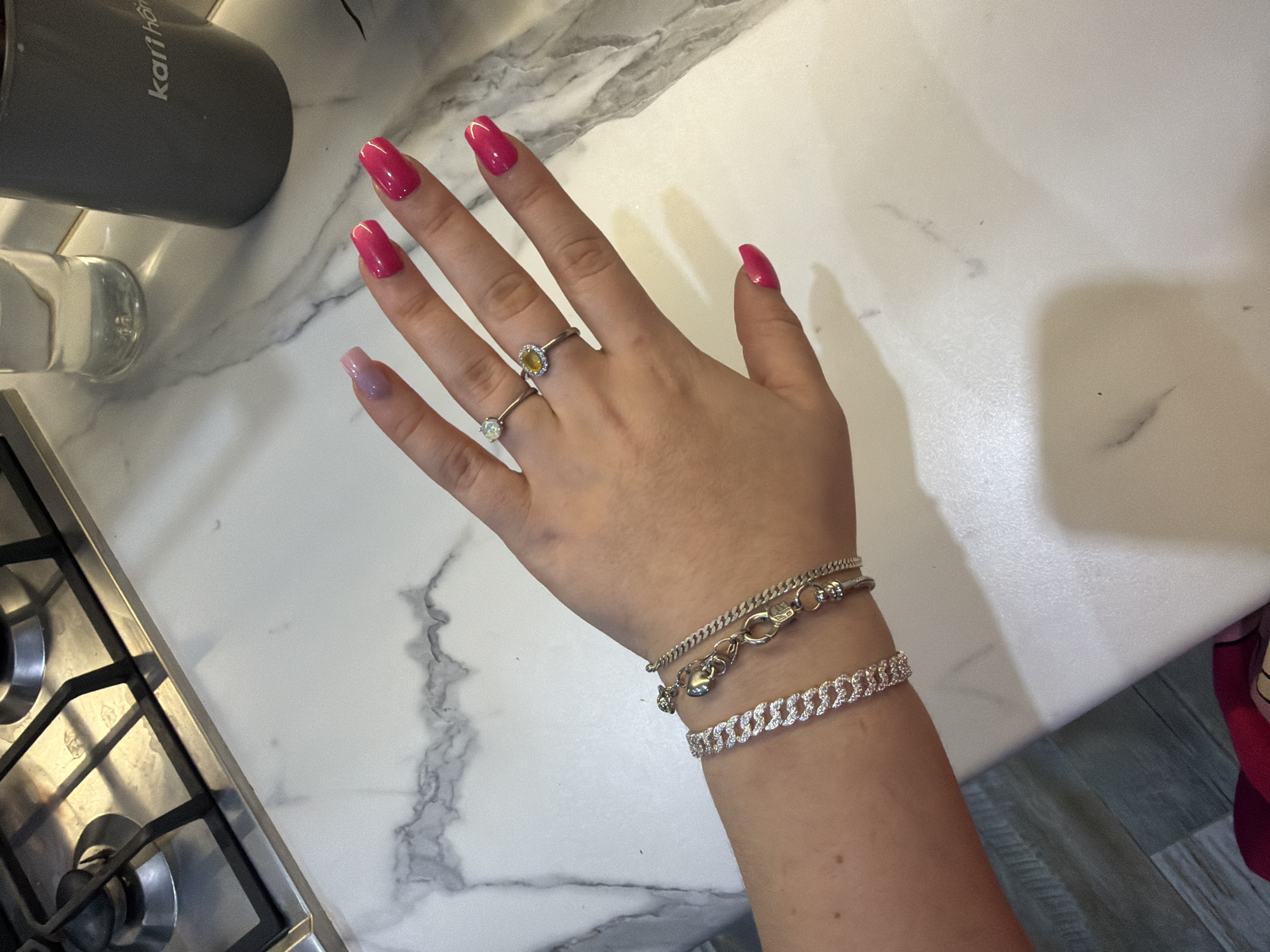 A close-up, high-angle shot features a human hand adorned with jewelry and brightly colored nails, positioned over a white marble-patterned countertop. The hand, likely female, displays vibrant fuchsia nail polish on four fingers (index, middle, ring, and thumb), while the pinky finger sports a lighter, gradient pink polish.
Two silver-toned rings are visible on the ring finger: one features a central oval-cut yellow stone surrounded by smaller clear stones, and another simple ring holds a single clear or light-colored oval stone. On the wrist, two silver-toned bracelets are worn. The upper bracelet is a delicate curb chain with a toggle clasp and a small, heart-shaped charm. Below it, a wider curb-link style bracelet is entirely encrusted with sparkling clear stones.
The background elements suggest a kitchen setting. To the upper left, a dark gray mug with the white text "kariho" or "karihome" is partially visible, next to a clear glass or bottle. In the lower-left quadrant, a metallic gas stovetop with black grates and burners is visible. The countertop features a white base with subtle gray marbling. The lighting is even, suggesting an indoor environment.