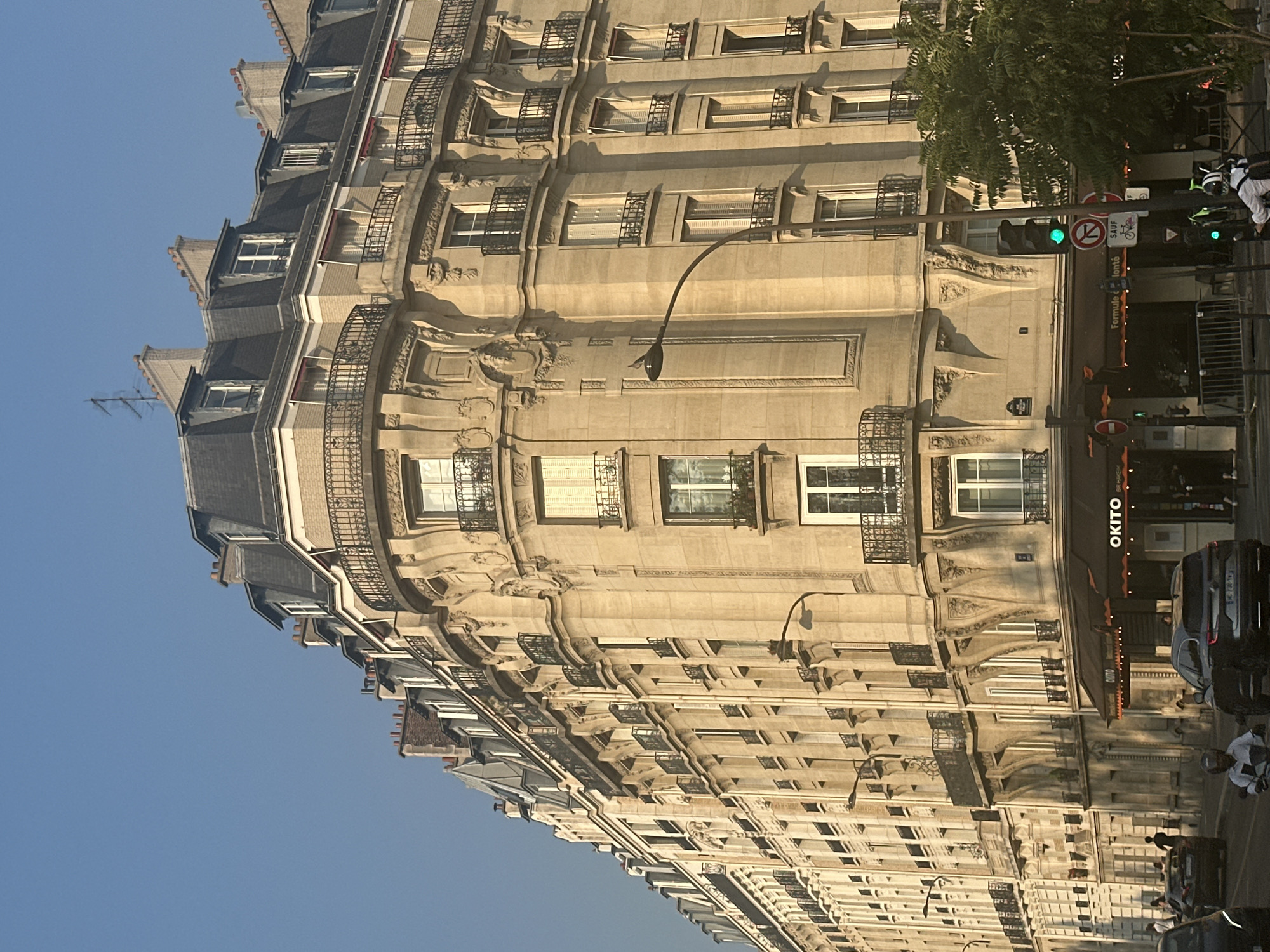 The image depicts a sunlit Parisian street scene dominated by a large, ornate Haussmannian-style building. The building, constructed of light-colored stone, features multiple floors with numerous windows, many adorned with wrought-iron balconies. The architecture exhibits classical detailing, including decorative carvings and moldings around the windows and cornices. The mansard roof is clad in dark slate with dormer windows.
At street level, the building houses a restaurant with an awning displaying the name "OKITO." Several cars are visible on the street, including a dark SUV in the foreground and a motorcycle with a rider. Pedestrians are also present, walking along the sidewalk and crossing the street. A traffic light with a green signal is visible. A mature tree with green foliage stands to the right of the frame, partially obscuring the building. The sky is clear and blue, indicating a sunny day. The overall impression is a typical urban streetscape in Paris, likely in the central or western arrondissements, known for their Haussmannian architecture.