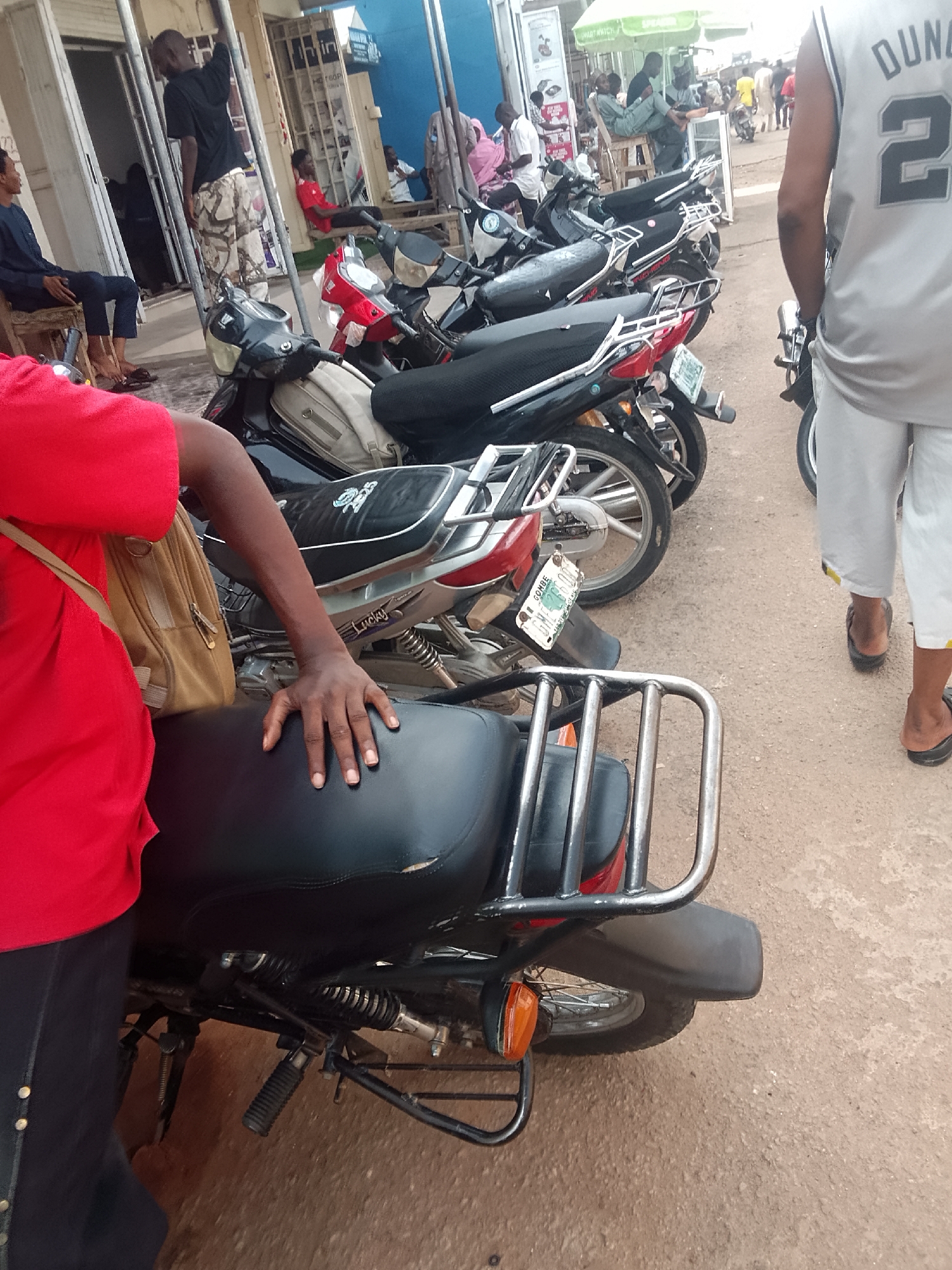 The image depicts a street scene in Gombe, Nigeria. A row of motorcycles is parked along the side of a paved road. In the foreground, a person in a red shirt and dark pants rests their hand on the seat of one of the motorcycles. Behind the motorcycles, people are seen standing and sitting along the street. One person is wearing a grey basketball jersey with the number 22 on the back. A building with various shops and advertisements lines the street in the background.