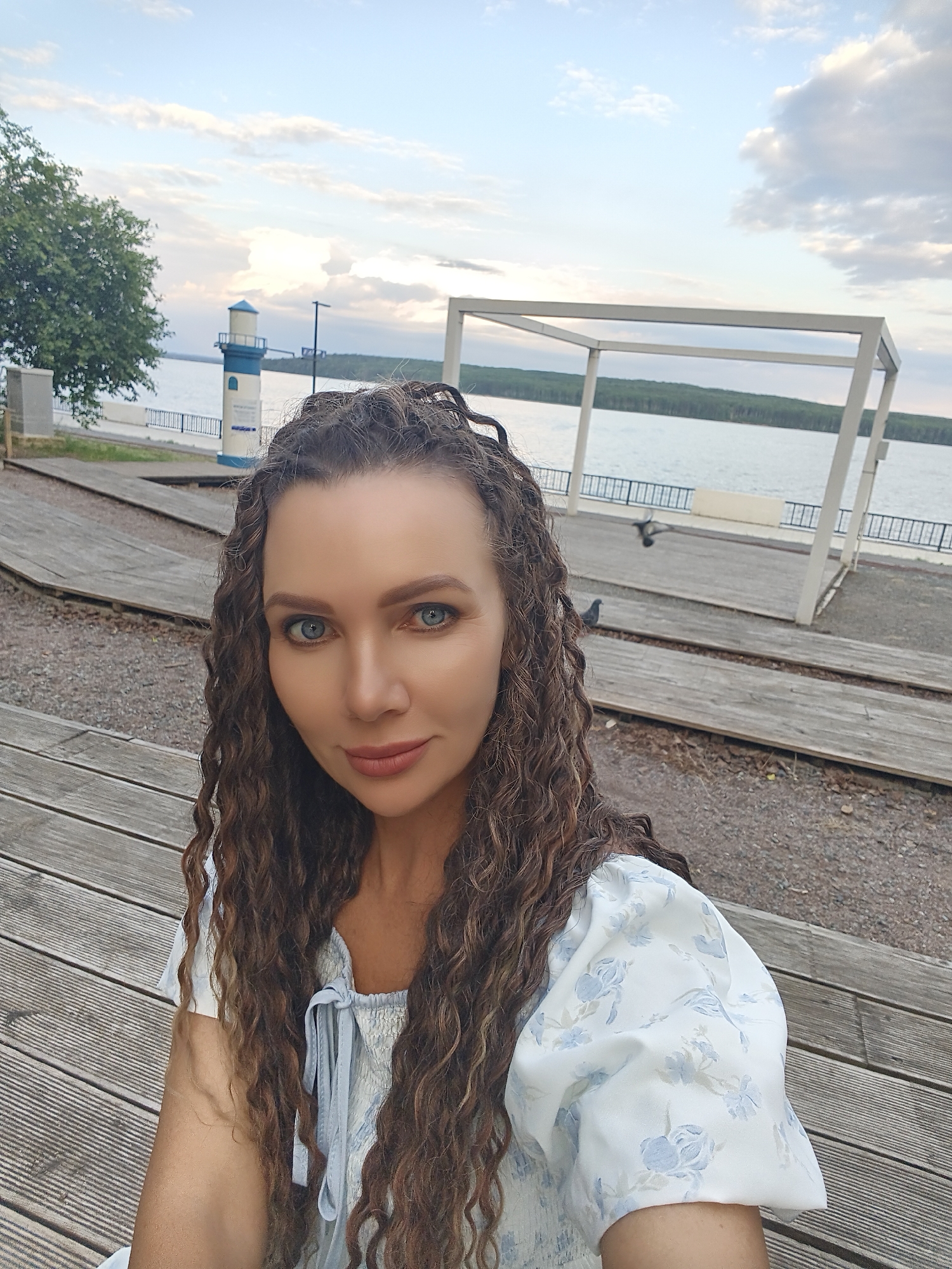 A woman with long, curly brown hair and blue eyes is taking a selfie outdoors. She is wearing a light blue and white floral print dress. The background shows a body of water, possibly a reservoir or river, with a lighthouse and a white gazebo structure. The ground is made of wooden planks and gravel. The sky is cloudy with a hint of sunset colors. This location is the embankment in the city of Chaikovsky, Russia, a popular place for recreation and enjoying the scenic views of the Kama River.