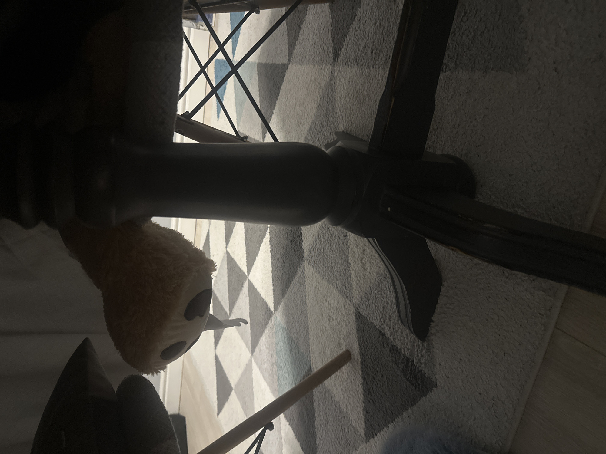 The image shows the underside of a table with a plush toy hanging from it. The toy appears to be a light brown color, possibly a teddy bear, and has two dark circular eyes visible. Its head is hanging down, with a light-colored string or thread dangling from its mouth area. The table has a dark, carved wooden pedestal base. The floor beneath the table is covered with a rug featuring a geometric pattern of light grey, dark grey, and white diamonds. Several chair legs are visible, one made of light wood and another with a black crisscross design. The scene appears to be indoors, possibly in a home environment. There is no indication of the city of Sofia, Bulgaria, within the image itself.