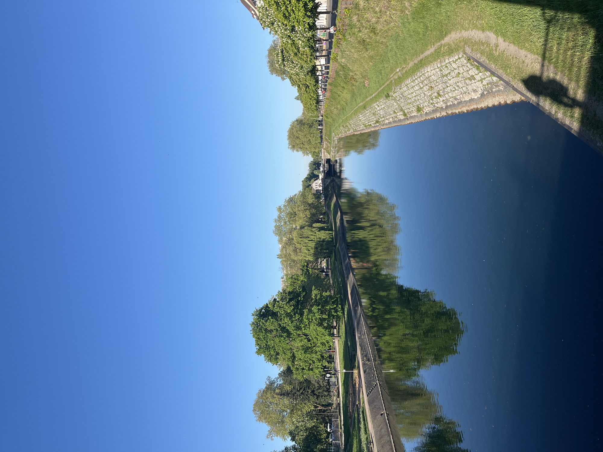 The image depicts a canal in Strasbourg, France, reflecting trees and the clear blue sky. A paved pathway runs along the canal's edge, bordered by green grass and stone embankments. The trees lining the canal create a reflection in the water, mirroring their lush foliage. The canal's water surface is mostly still, enhancing the clarity of the reflection. The shadow of a bridge or overhanging structure is visible on the right side of the canal.