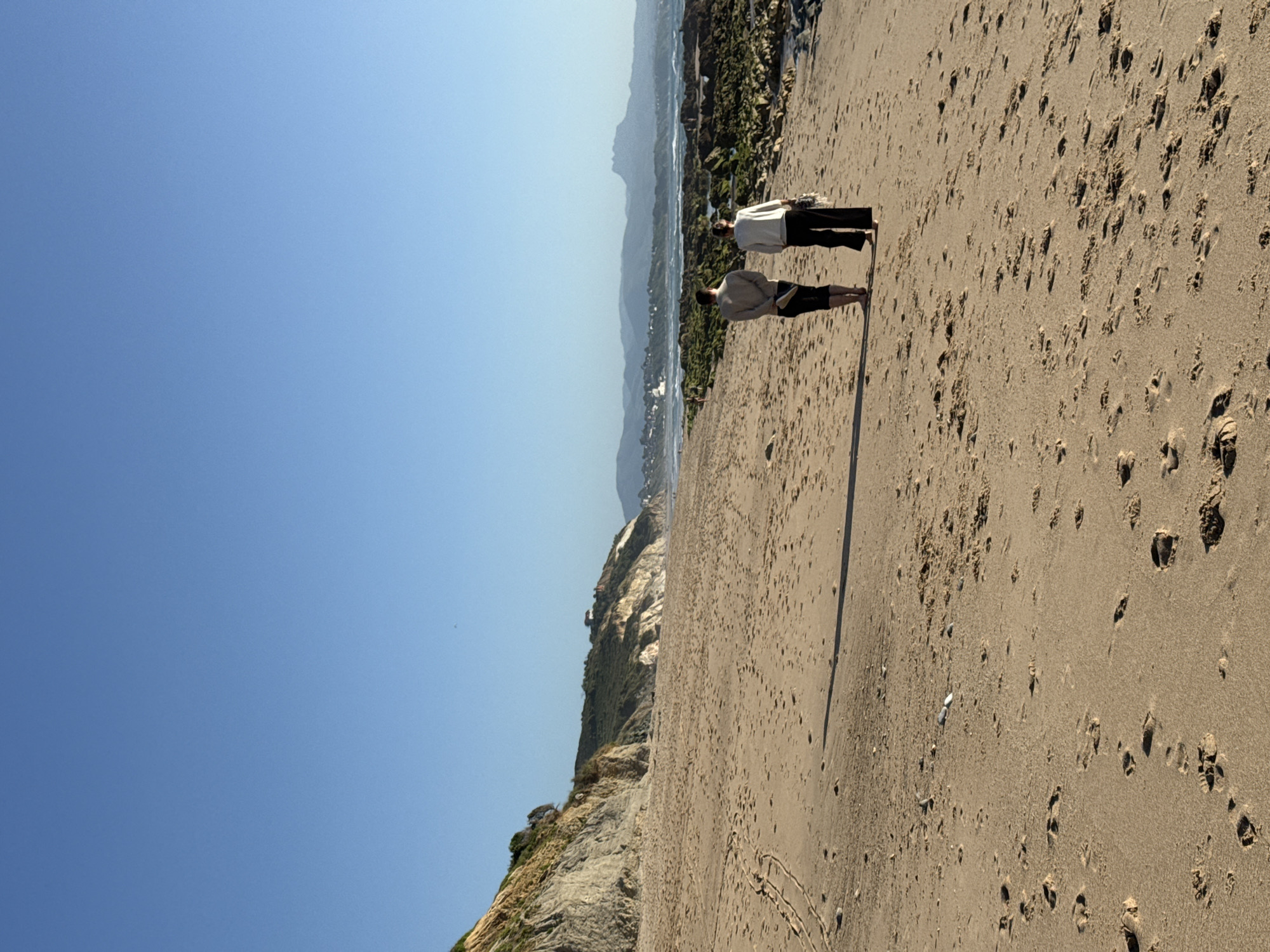 Two individuals are walking away from the camera on a sandy beach under a clear blue sky. The man on the left is wearing a light-colored long-sleeved shirt and dark pants, holding a pair of shoes in his left hand. The woman on the right is wearing a white long-sleeved shirt and dark pants, carrying a folded item in her right hand. The beach is covered in footprints and small pebbles. In the background, to the left, is a steep, rocky cliff with vegetation. Further in the distance, a town is visible along the coastline, nestled among green hills. The ocean stretches out to the horizon, with waves breaking against rocky outcrops covered in green algae. The overall scene suggests a peaceful walk along the coast. This scene is likely the Plage de la Milady in Biarritz, France, known for its expansive sandy areas and cliffside views.
