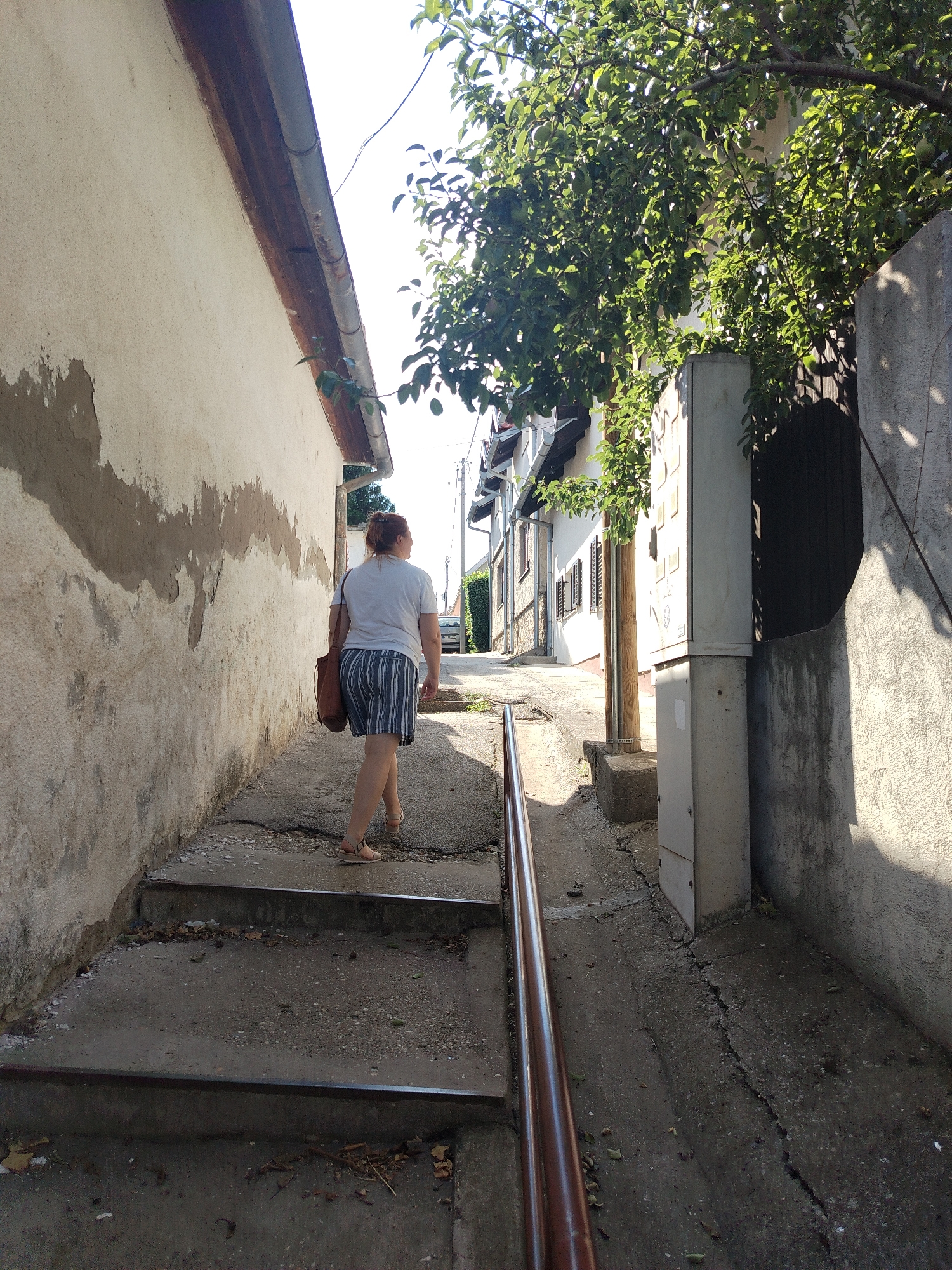 A woman with auburn hair pulled back is walking up a set of concrete stairs in an alleyway. She is wearing a white t-shirt and blue and white striped shorts, with tan sandals. A brown leather shoulder bag is slung over her right shoulder. The alleyway has rough, textured walls on both sides. The wall on the left is painted a light beige with patches of darker, exposed plaster. The wall on the right is rough, grey concrete and appears to have a dark wooden gate or door set into it. Above the alley, green leaves and branches from a tree are visible, casting shadows on the scene. In the distance, at the top of the alley, houses and a car are visible, suggesting a residential area. The ground of the alley is cracked concrete. The scene appears to be in Sremski Karlovci Municipality, Serbia, based on the context provided.