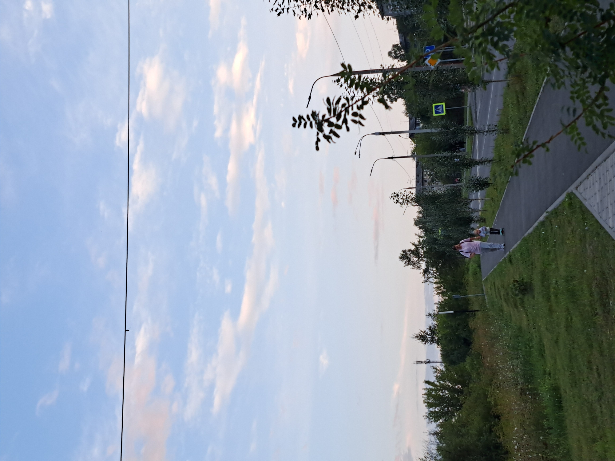 The image depicts a street scene with a sidewalk and a road. On the sidewalk, a woman in a pink shirt and grey pants walks alongside a child on a scooter. The child is wearing a jacket and pants. They are walking away from the viewer. The sidewalk is bordered by a grassy area with some wildflowers. Along the road, there are several young trees lining the street. Streetlights are visible, and there are buildings in the background. A pedestrian crossing sign is also visible on the right side of the road. The sky is cloudy with patches of blue. The presence of streetlights, apartment buildings, and the general urban infrastructure suggest this is likely a residential or commercial area within a city. Without further information, it's impossible to definitively pinpoint the exact location within Irkutsk, Russia, but the scene is characteristic of a typical Russian urban environment.