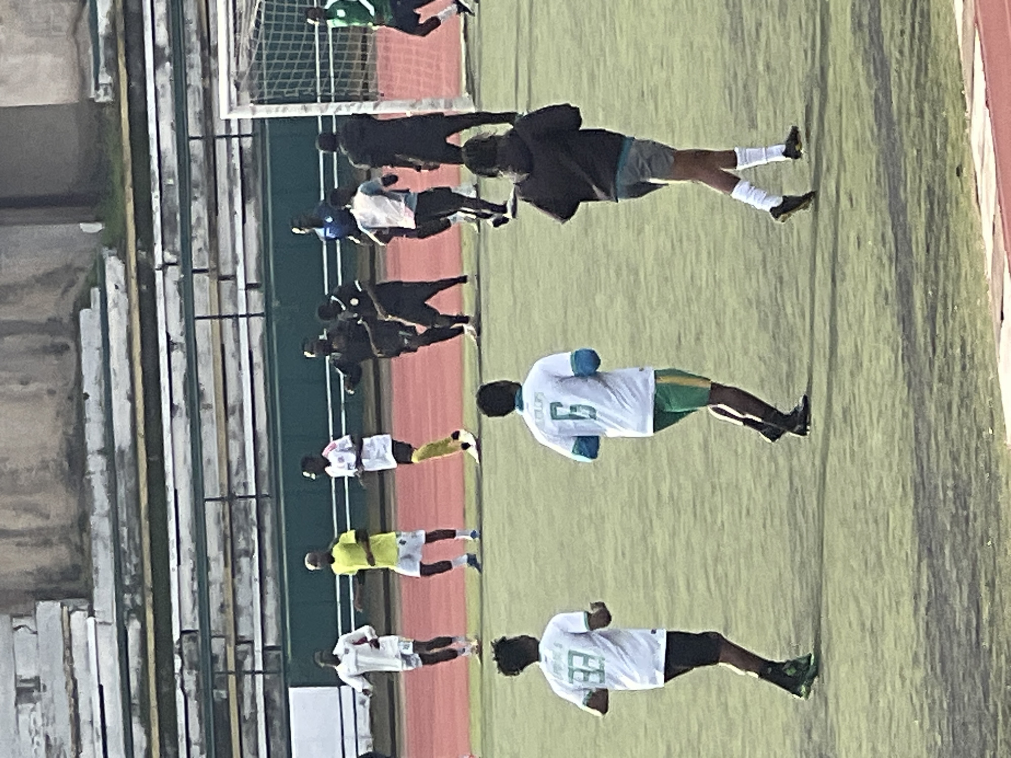 The image shows a group of male soccer players on a green field at the New Stadium in Nigeria. The players are in various positions, with some running and others standing. The stadium appears to be mostly empty, with rows of empty seats visible in the background. The players are wearing different colored uniforms, including white, yellow, black, and green. Some players have numbers on their jerseys. A soccer goal is visible in the background on the right side of the image. The lighting suggests it is daytime, with the sun casting shadows on the field. The overall scene depicts a soccer practice or warm-up session.