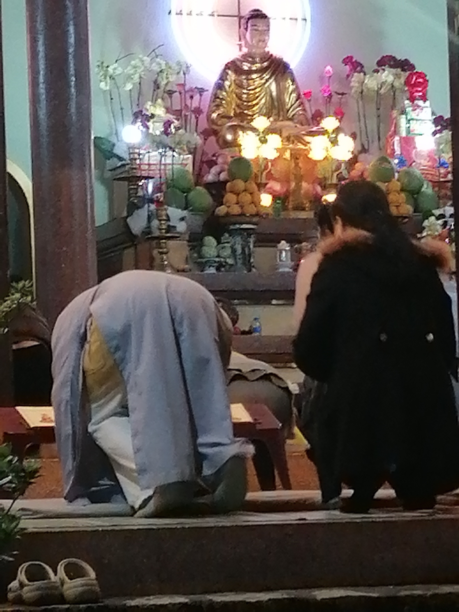 The image depicts a scene inside a Buddhist temple in Da Lat, Vietnam. In the background, a golden statue of Buddha is seated, adorned with a flowing robe. The statue is surrounded by an altar decorated with offerings of fruit, flowers, and lit candles. Two individuals are visible in the foreground. On the left, a person in a light blue robe is kneeling with their head bowed. On the right, another person is seen from behind, wearing a dark coat. The environment suggests a place of worship, with architectural elements of a temple visible, including pillars and ornate decorations. The lighting appears dim, characteristic of indoor temple settings.