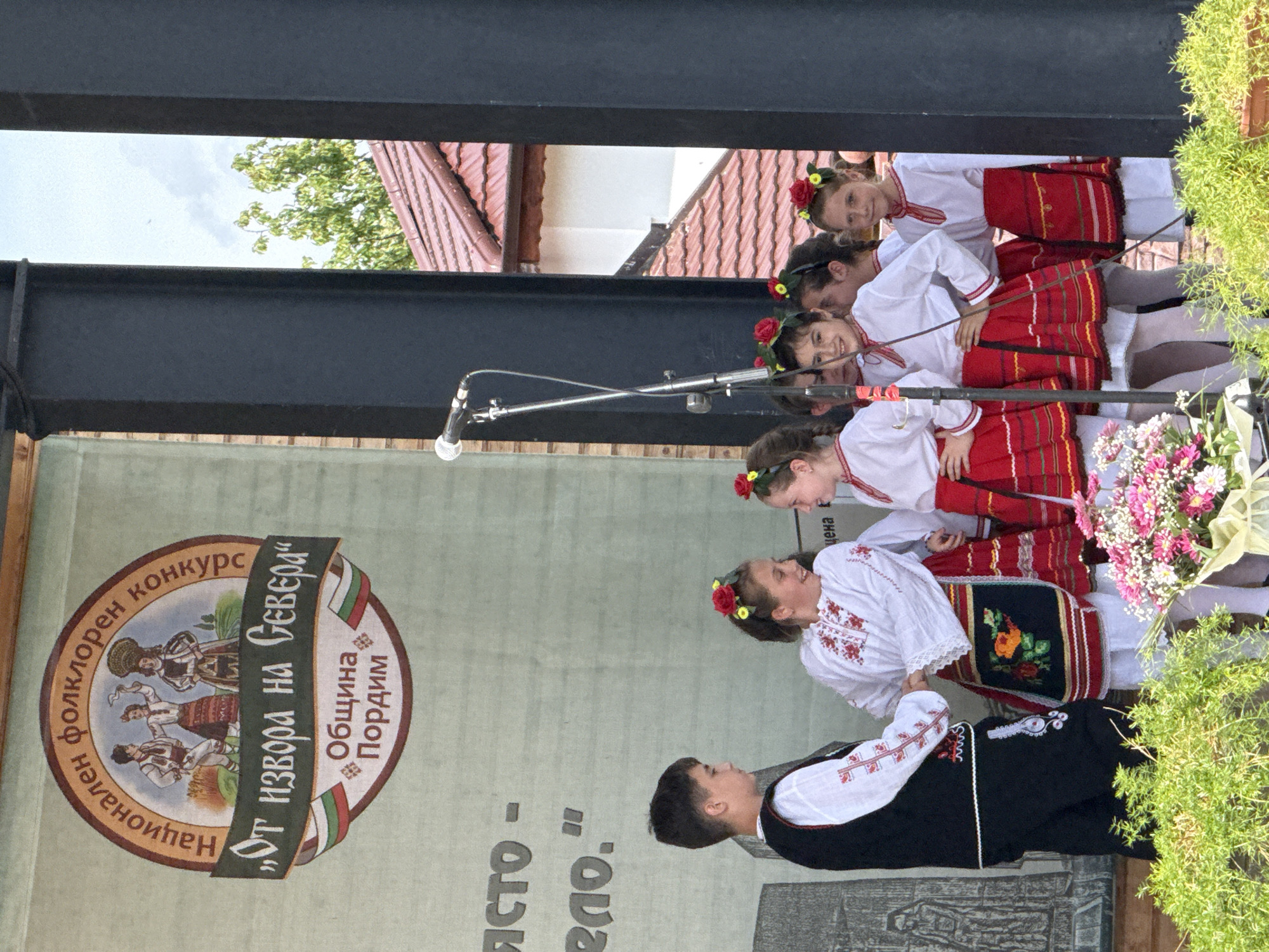 This image captures a performance on an outdoor stage in Pordim, Bulgaria. Several young people, dressed in traditional Bulgarian folk costumes, are the main subjects. On the left, a boy in a black vest over a white embroidered shirt and black pants stands facing a group of girls. The girls are wearing white embroidered blouses with red trim and red skirts with black and white patterned aprons. They have floral headpieces. Some of the girls are smiling and interacting with each other, while others are looking forward.
Behind the performers, a large banner displays a circular emblem with text in Bulgarian. The emblem features figures in traditional attire and the text reads "Национален фолклорен конкурс" (National Folklore Competition) at the top, and "„От извора на Севера“" (From the springs of the North) below. Further down, it says "Община Пордим" (Municipality of Pordim), indicating the location of the event. There are also Bulgarian flags incorporated into the design.
To the left of the banner, some text is visible, reading "ЯСТО -" and "ело." which might be part of a larger backdrop. A microphone on a stand is positioned in the center of the stage, suggesting a musical or vocal performance.
The background reveals elements of the environment. Through a gap between black structural beams, a glimpse of the sky, trees, and a red-tiled roof of a building can be seen. In the foreground, lush green foliage and a bouquet of pink and white flowers are arranged, adding to the festive atmosphere. The overall scene depicts a cultural event celebrating Bulgarian folklore in the town of Pordim.