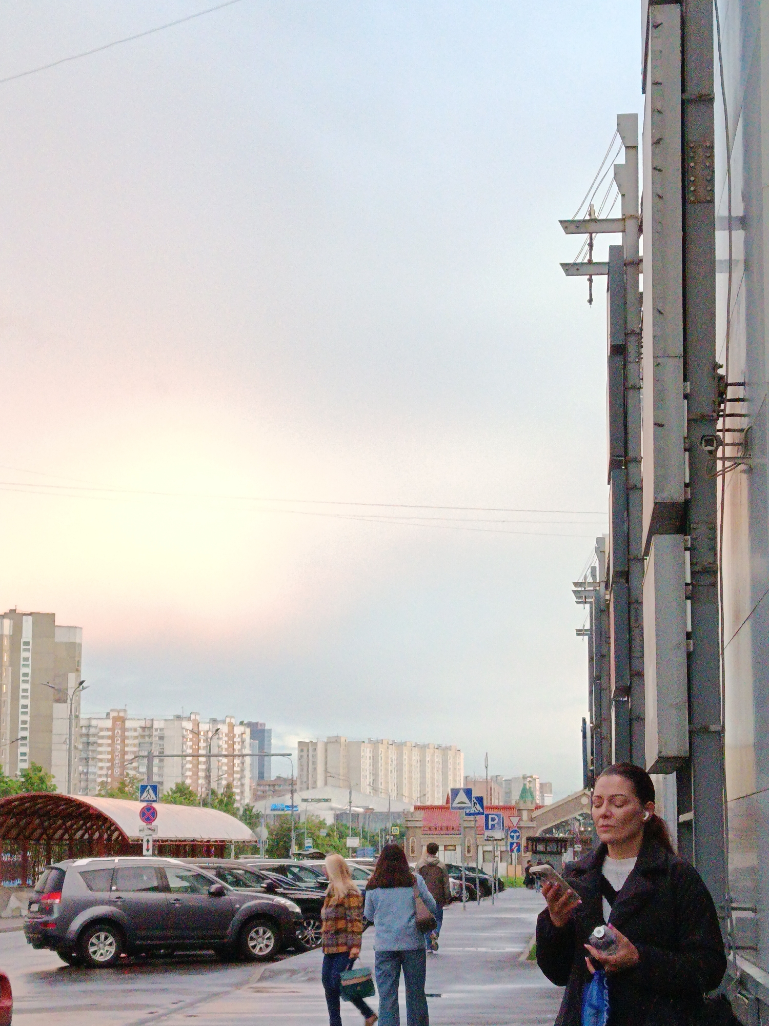 The image captures a street scene in Moscow, Russia, on an overcast day. The backdrop features apartment buildings of varying heights, typical of Moscow's urban landscape. On the left, a covered walkway and traffic signs are visible amidst parked cars, suggesting a busy area.
A woman stands on the sidewalk to the right, appearing engrossed with her mobile phone. She holds a water bottle and a blue bag, while another woman and a man walk ahead of her on the sidewalk. The right side of the frame is dominated by a tall, industrial-looking building facade, possibly part of a shopping center or office complex. The overall atmosphere suggests a typical urban setting during a cloudy day in Moscow.