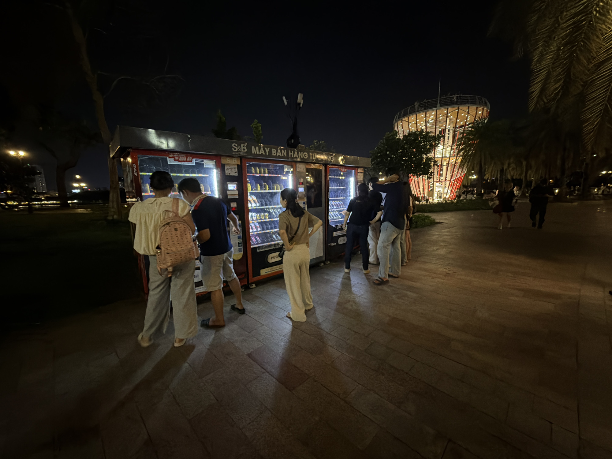 This nighttime image depicts a row of automatic vending machines situated along a paved walkway in Thủ Đức, Vietnam. The vending machines are illuminated, displaying a variety of packaged beverages and snacks. A canopy above the machines bears the Vietnamese text "S&B MÁY BÁN HÀNG TỰ ĐỘNG," indicating "S&B Automatic Vending Machine."
Approximately six to eight individuals are gathered around the vending machines. From left to right: a person in a light-colored shirt and wide-leg pants with a pink backpack stands facing a machine; next, a person in a dark blue polo shirt and light shorts interacts with an adjacent machine; further right, a person in a brown top and light pants stands facing another machine with hands on hips. Other individuals are partially visible further down the line of machines, including one in a dark t-shirt and light pants, and another in a dark top and light jeans.
In the midground to the right, a distinctive, multi-story, illuminated building with a conical or cylindrical shape and an external spiral walkway is prominent. Palm trees are visible throughout the scene, particularly on the right, where their fronds are silhouetted against the dark sky. Other trees and distant artificial lights are discernible on the left and in the background. The ground is a dark, patterned paved surface. Additional pedestrians are visible walking in the vicinity of the illuminated building in the background.