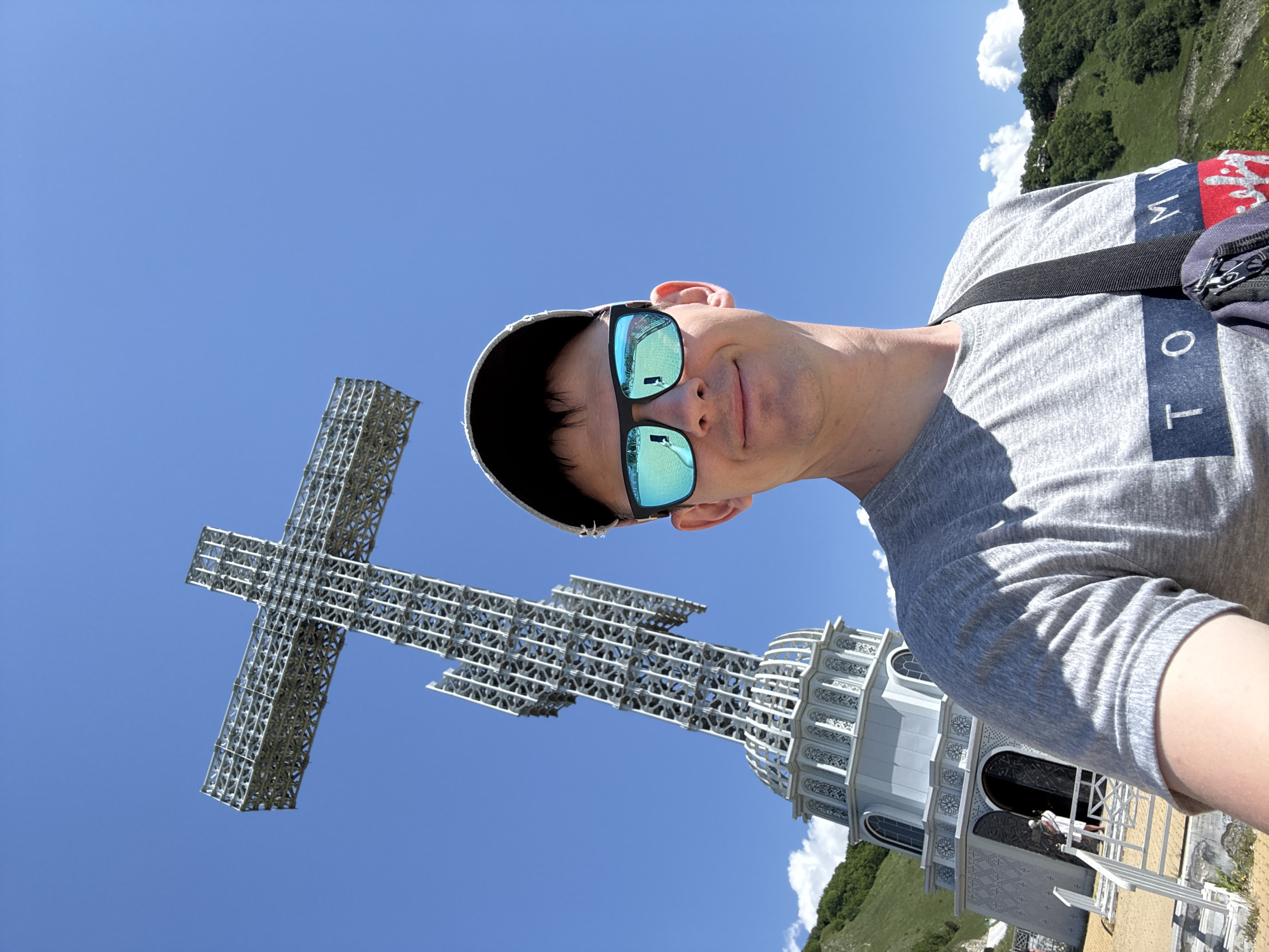 This image is a bright, sunlit selfie taken by a man at an outdoor landmark. The man, positioned in the foreground, is looking directly at the camera with a subtle smile. He wears a grey t-shirt, distinctive blue mirrored sunglasses, and a light-colored cap. A black shoulder bag strap crosses his chest. Visible text on his t-shirt includes "M Y" and "T O", suggesting the brand "TOMMY".
In the midground, a towering metallic cross monument, constructed from an intricate lattice or truss framework, stands prominently against the sky. Below and to the right of the cross, a grand, light-colored building with a domed or multi-tiered roof and decorative arched windows is visible. Several indistinct people can be seen near the entrance of this building.
The scene is set under a clear, vibrant blue sky dotted with a few scattered white clouds. Lush green, hilly or mountainous terrain, covered with trees, forms the distant background, indicating an elevated location. The ground near the building appears to be an accessible pathway or plaza.
The bright sunshine indicates it is daytime, likely mid-morning or early afternoon, with clear weather. The man appears to be a tourist or visitor enjoying the panoramic views at this significant site. The location is Gelendzhik, Russia, and the cross is likely the Poklonny Cross, a notable landmark in the area.