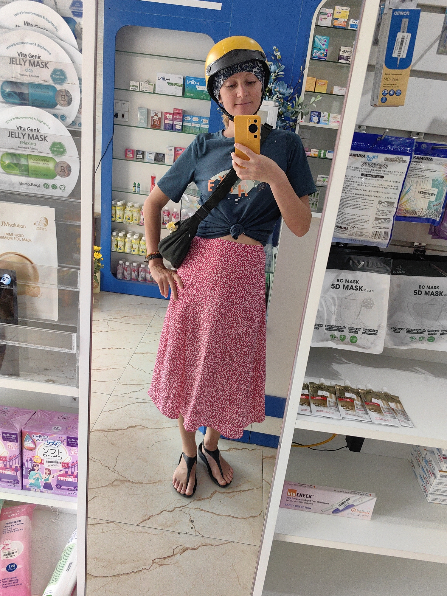 A waist-up shot shows a woman in a pharmacy in Bai Chay Ward, Vietnam, as seen through the mirrored door of a product display cabinet. The woman is wearing a yellow helmet over a patterned headscarf, a teal T-shirt tied at the waist, a knee-length pink and white patterned skirt, and black sandals. A black crossbody bag hangs over her shoulder. She is holding a yellow smartphone to take a selfie.
The shelves behind the woman display a variety of products including face masks, thermometers, and hygiene products. The floor is tiled.
The shelves in the cabinet on the left contain cosmetics and facial treatments. The shelves in the cabinet on the right contain face masks, oral hygiene products, hair treatments, and pregnancy tests.