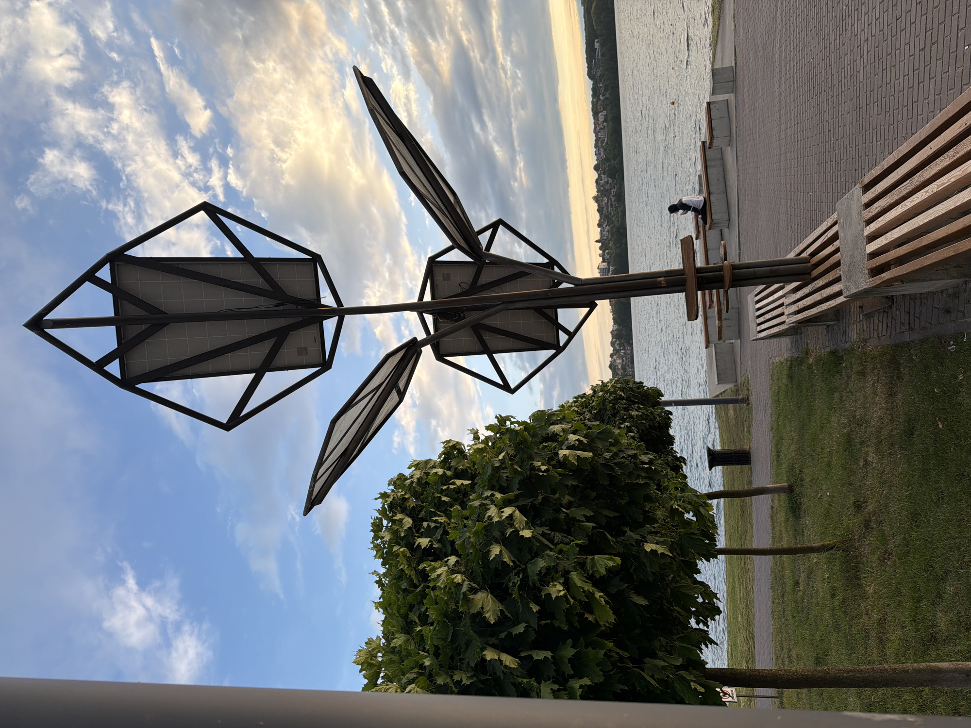 The image depicts a waterfront area in Ternopil, Ukraine, likely along the Ternopil Reservoir. In the foreground, a modern-designed lamppost with solar panels is prominent. To the left, manicured trees with full green foliage line a grassy area. A paved walkway runs alongside the water, furnished with wooden benches. In the middle ground, a person is seated on a bench, looking out at the water. The background features a distant cityscape with buildings and lush greenery bordering the water, under a sky with scattered clouds at dusk.