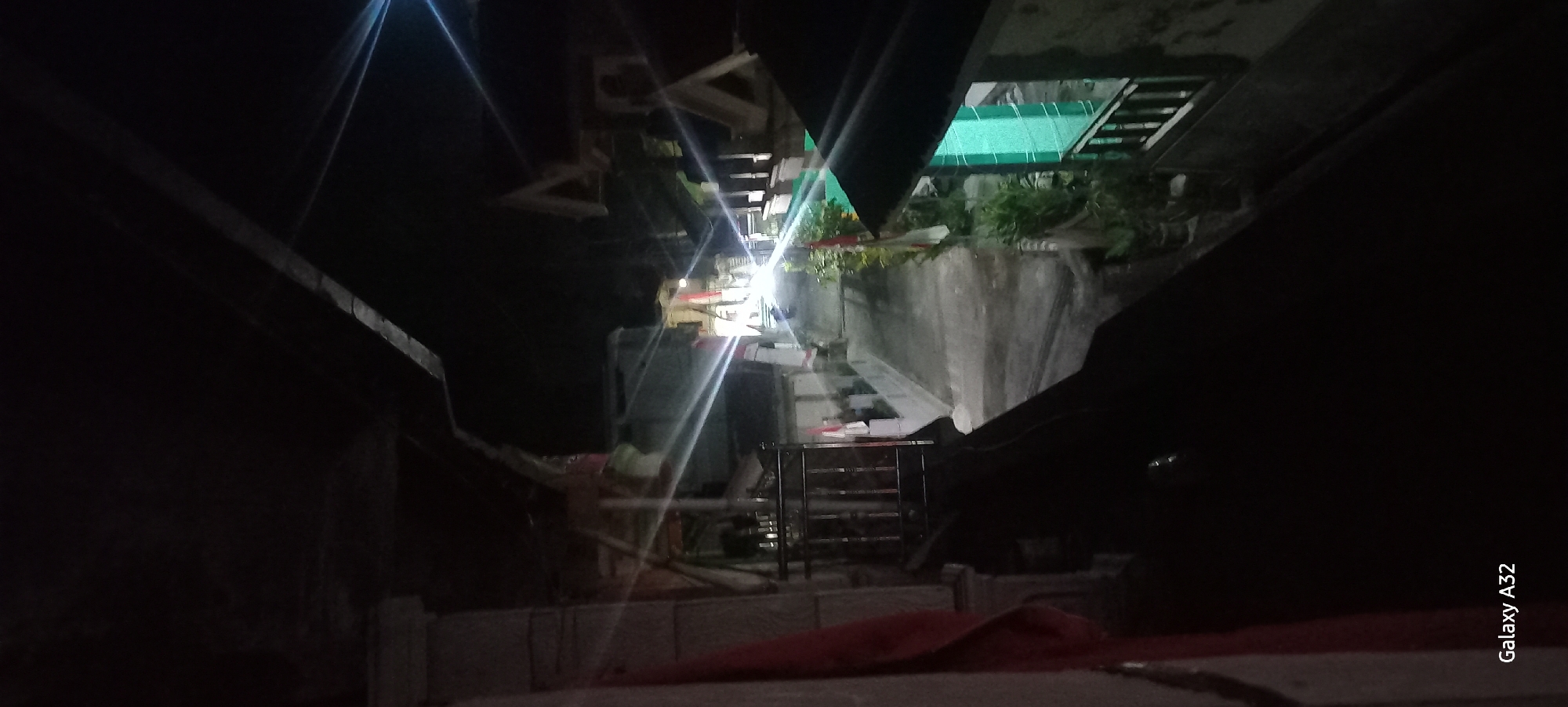 This is a nighttime photograph taken from a high vantage point, looking down a street in Plumpung, Indonesia. The street is illuminated by bright, possibly artificial, lights that create strong glare and lens flares throughout the image. On either side of the street are buildings and houses with visible roofing and window structures. Some of these buildings have decorative elements, including what appear to be flags mounted on poles, possibly indicating a festive occasion or celebration. There are also plants in pots along the side of the road. The street surface itself is visible and appears to be paved. The foreground of the image is dark, with a shadowed area obscuring details, suggesting the photographer is looking from a covered balcony or elevated structure. The bottom left corner of the image displays the text "Galaxy A32", indicating the device used to capture the photograph.