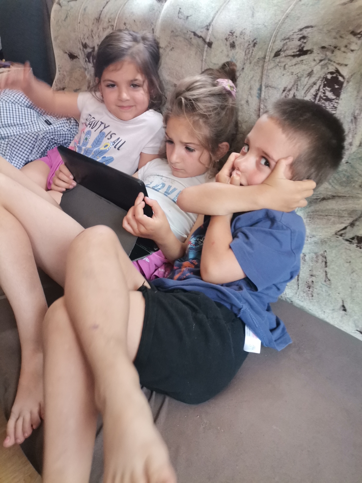This image, captured in Obrochishte, Bulgaria, presents three children seated closely together on a patterned couch with a dark grey base cushion.
On the left, a female child, estimated to be between 5 and 7 years old, has dark hair and wears a white T-shirt with the phrase "BEAUTY IS ALL" printed on the front, along with pink shorts and a plaid shirt partially visible on her left side. She looks directly at the viewer with a slight smile, her left arm raised and bent at the elbow.
In the center, another female child, also estimated to be between 5 and 7 years old, has light brown hair secured with a pink hair tie. She wears a white T-shirt and pink shorts, and is intently focused on a black tablet, held in a dark grey case, positioned on her lap. Her hands are on the tablet's screen.
To the right, a male child, estimated to be between 5 and 7 years old, has short dark hair and wears a blue T-shirt and black shorts. His left hand is positioned near his mouth and nose, while his right arm extends around the head area of the central child. He looks towards the upper right of the frame with a wide-eyed expression. A small, dark mark is visible on his right shin.
The children occupy the front of the image, with the patterned backrest of the couch forming the primary background. The setting appears to be an indoor environment, illuminated by general ambient light.