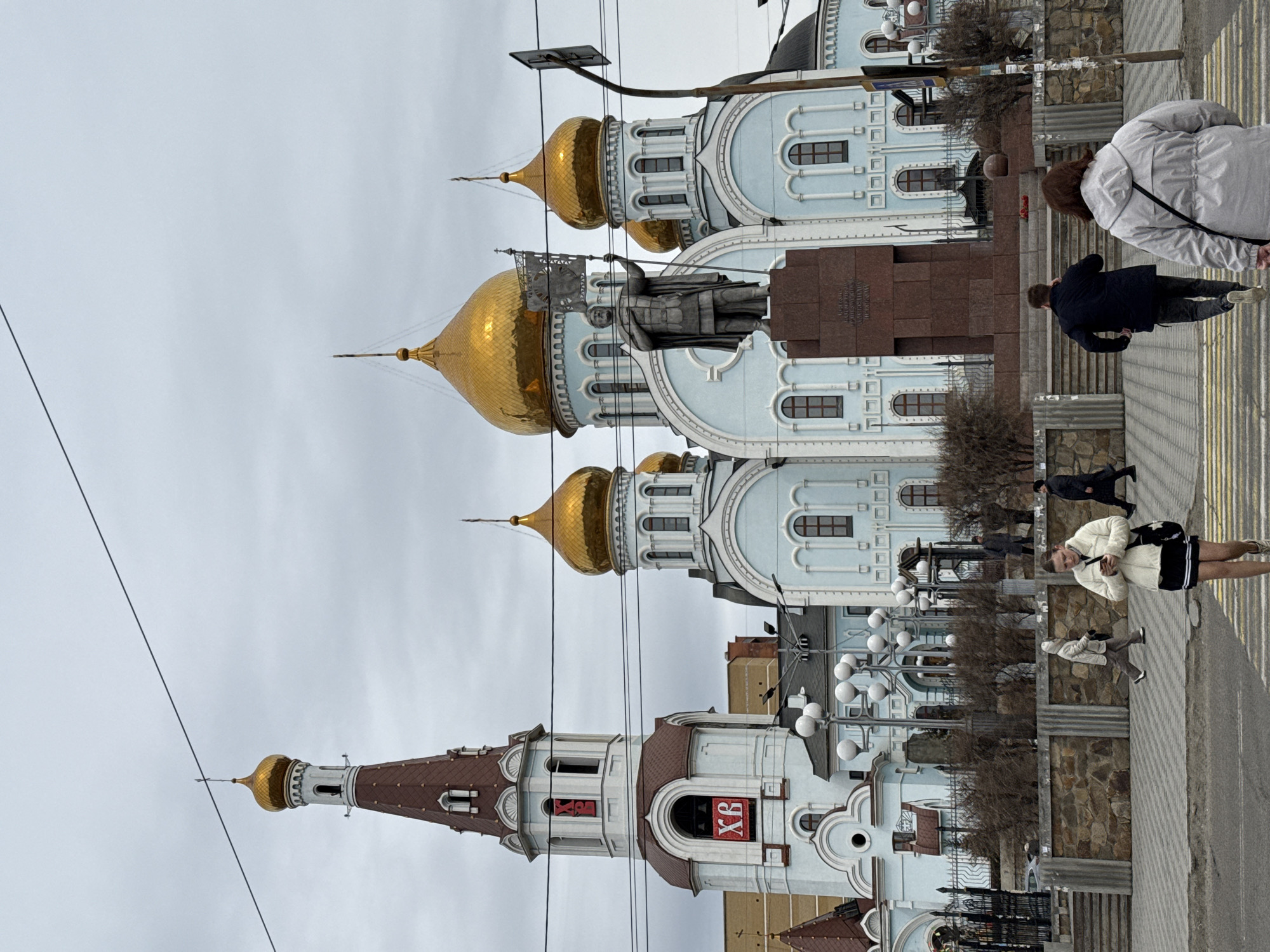 The image captures a view of the Pokrovskiy Cathedral in Vladivostok, Russia. The cathedral features multiple gold domes and white-and-blue facades. In front of the cathedral is a statue on a tall pedestal. Pedestrians are walking on the sidewalk. Overhead, power lines are visible. The sky is overcast.