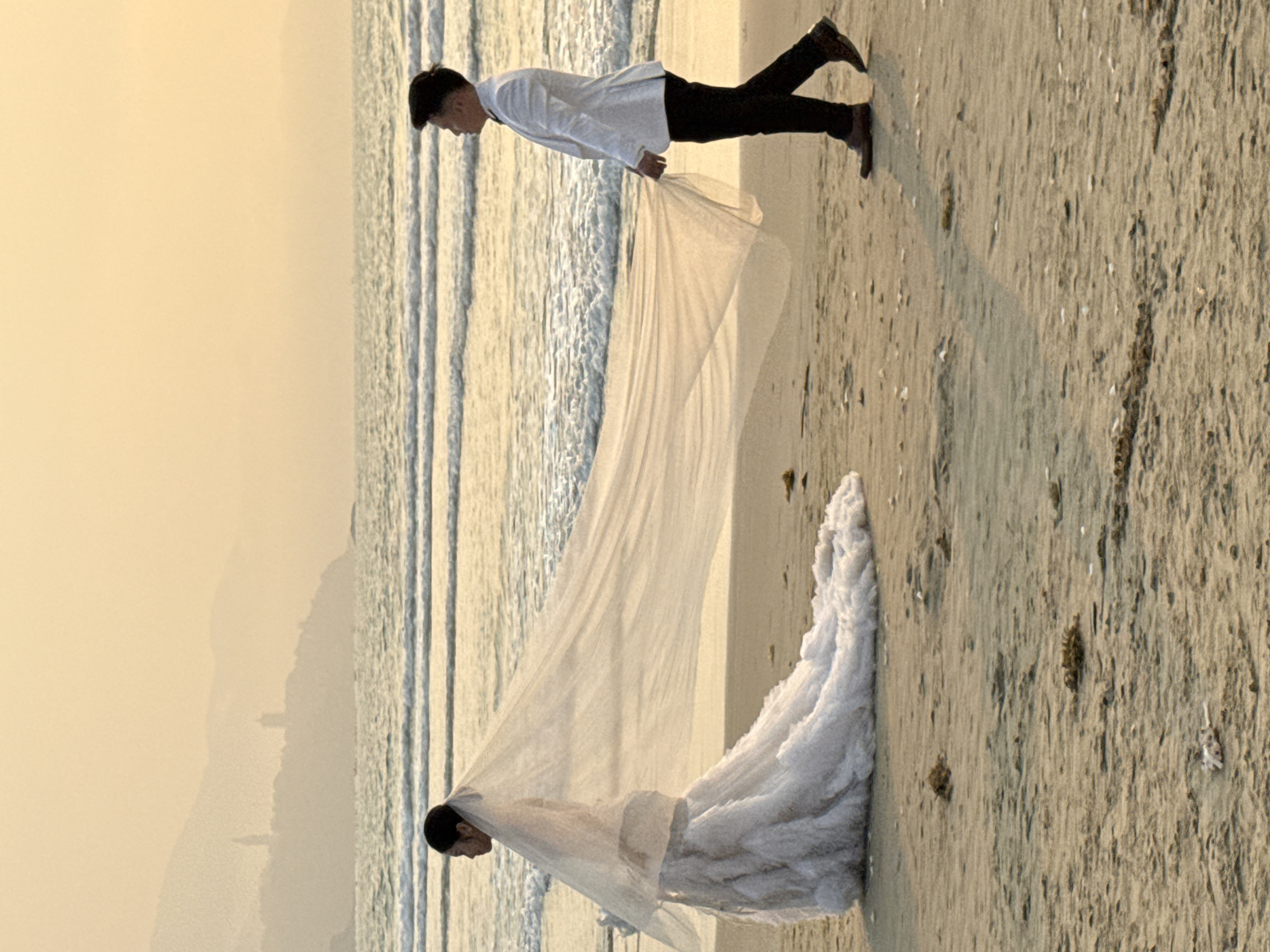 On a beach in Đà Nẵng, Vietnam, a groom in a white shirt and dark pants holds the flowing veil of a bride in a wedding dress. The couple stands near the shoreline, with the sea to their left and sandy beach to their right. The sky is overcast.