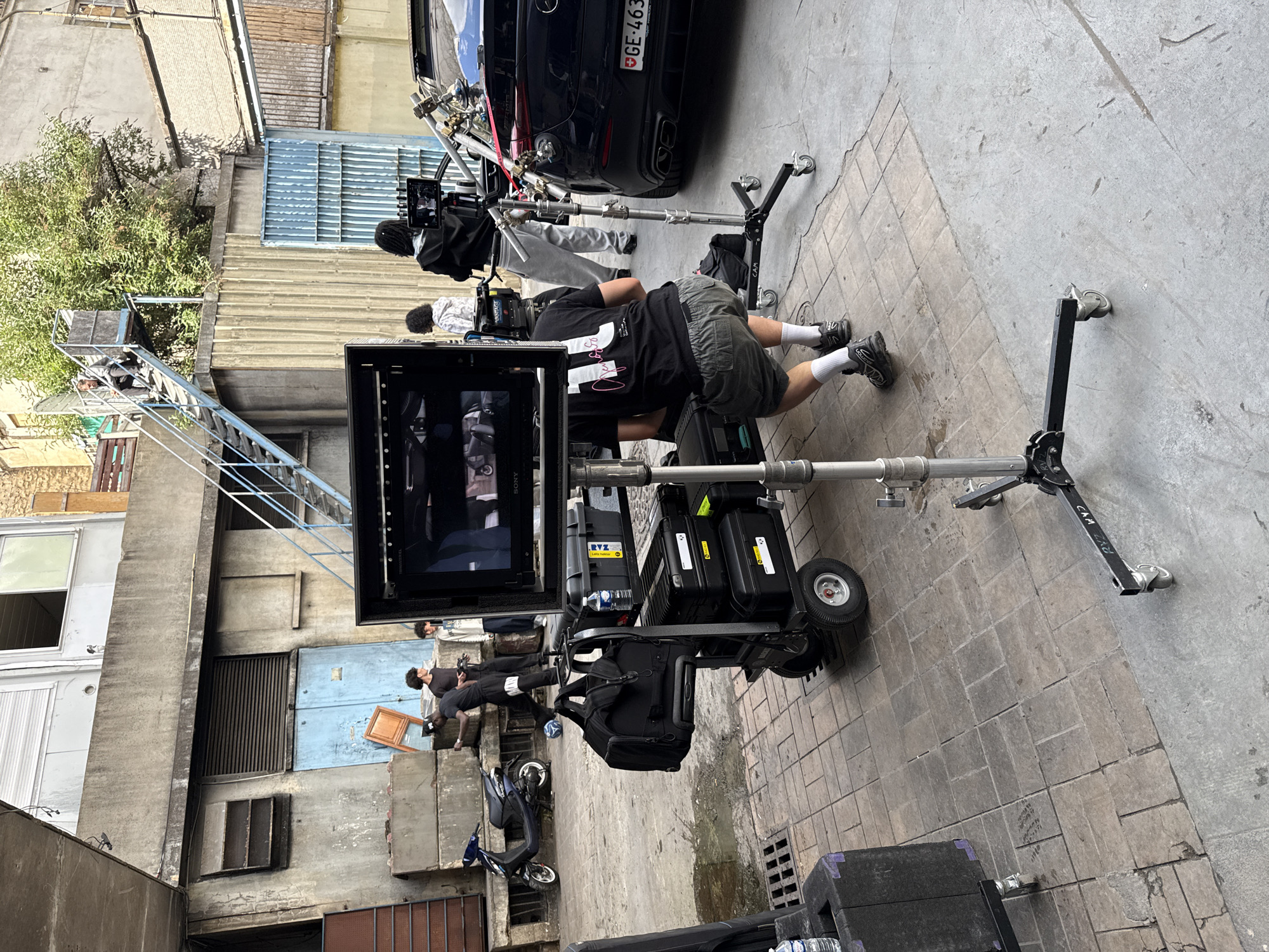 The image captures a film shoot in an urban alleyway, likely Ivry-sur-Seine, France, based on the car's license plate. A black car is partially visible in the background, alongside film equipment. A crew of people is present, including a person lying on the ground near the camera monitor, and others operating film equipment. The environment is a mix of concrete buildings with windows, doors, and a metal staircase. A blue scooter is also parked in the alley. The lighting suggests it's daytime.