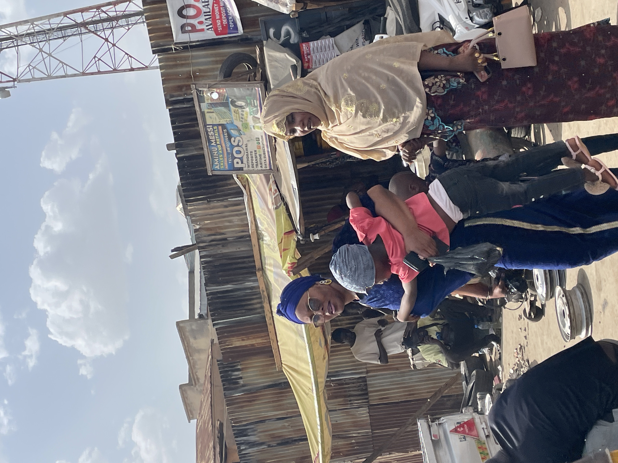 This image, captured in Jos, Nigeria, presents a vibrant outdoor street or market scene under a bright, partly cloudy sky. The setting features informal market stalls or buildings constructed with corrugated metal sheets.
Several people are visible, including two women prominent in the foreground. One woman, wearing a light-colored hijab with gold embellishments, looks towards the viewer while holding a small, light-colored purse. Adjacent to her, another woman in a blue headscarf and sunglasses also looks at the camera, holding a child in a pink top who is preoccupied with a dark object, possibly a phone. Other individuals are partially visible in the background, contributing to the bustling atmosphere.
The scene is rich with contextual details, including various signs advertising services common in Nigerian markets. A prominent sign reads "AMINU MESSI SALES AND RETAILS" with a phone number "TELL: 08103366615". Below this, "POS" (Point of Sale) services are listed, including "WITHDRAWAL", "DEPOSITS", "TRANSFER", "AIRTIME", and "DATA SUBSCRIPTION". Another partially visible sign repeats "P.O." and offers "AVAILABLE" services like "SEND MONEY", "DEPOSIT MO" (Deposit Money), and "OPEN ACC" (Open Account).
In the upper left, a large red and white metal tower structure, possibly a telecommunications mast, rises against the sky. The ground shows typical street clutter, including discarded items and vehicle wheels. The time of day is clearly daylight, indicated by the bright illumination. The image captures a lively and typical commercial interaction in a Nigerian urban environment.