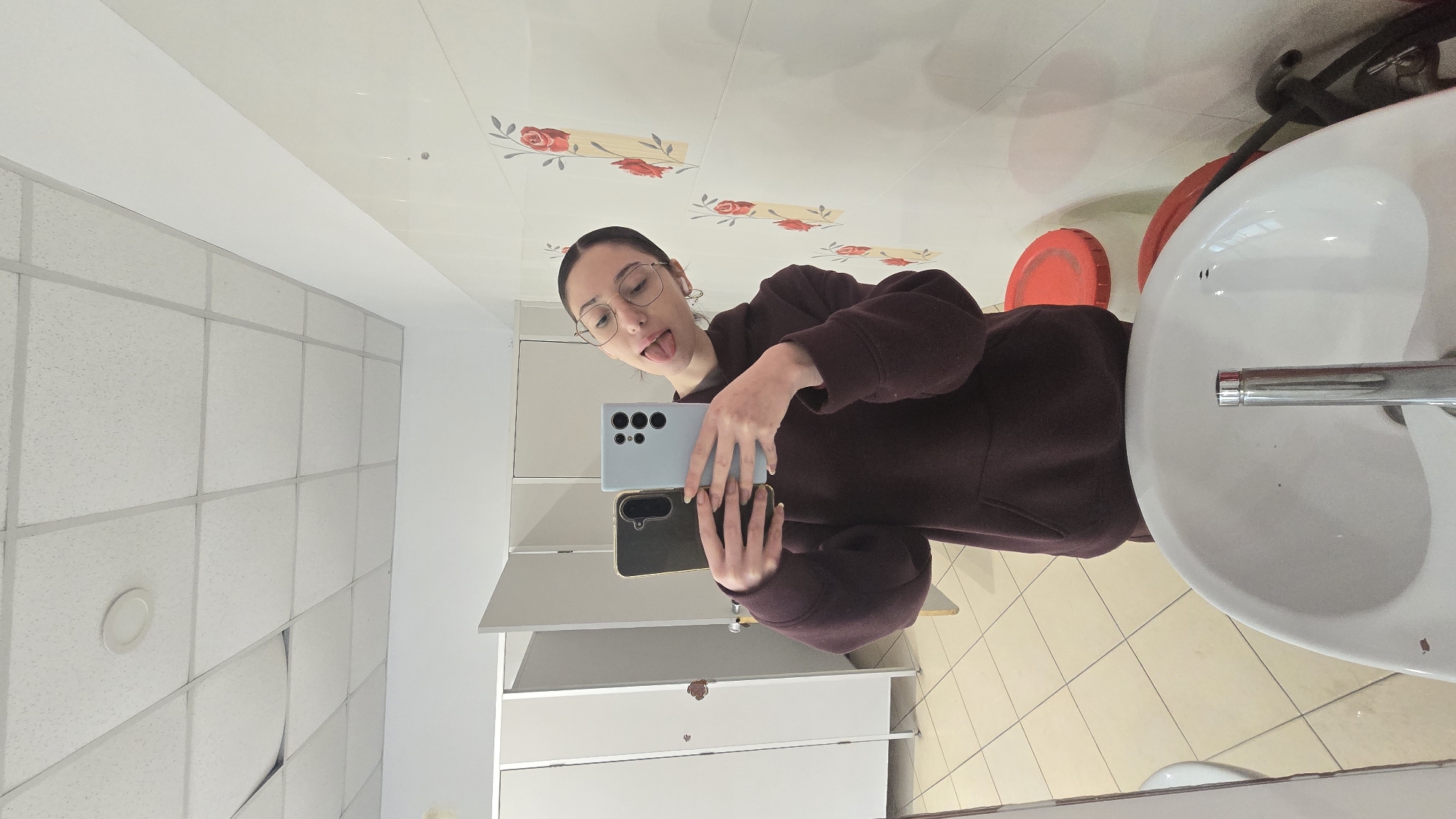 A mirror selfie in a bathroom of an unknown location in Stara Zagora, Bulgaria. A young woman with glasses and a brown sweatshirt holds two smartphones, one on top of the other, and sticks her tongue out toward the camera. Overhead is a ceiling with a pattern of flowers. To the right is a sink and the floor is tiled. To the left, bathroom partitions.