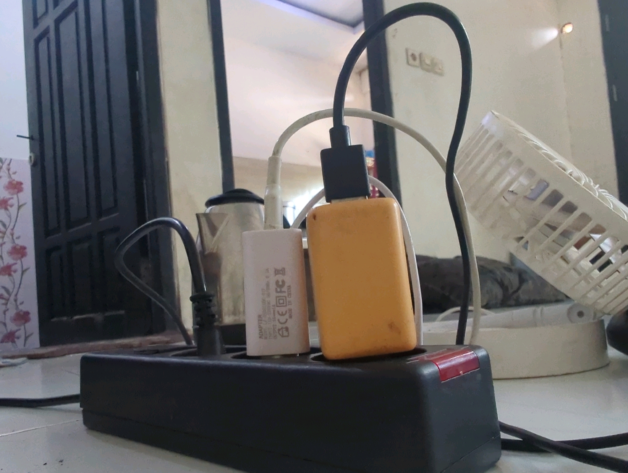 A low-angle shot captures a black power strip on a tiled floor, overloaded with various charging devices. Two white and orange charging adapters are plugged into the power strip, connected to black and white cables. A coiled black cable extends from the power strip, disappearing out of frame. In the background, a dark door with vertical panels is visible on the left. To the right of the door, a white wall with two electrical outlets and a small, circular light fixture illuminates the scene. Further to the right, a white electric fan with a perforated cage is tilted on its base, partially obscuring a gray, fuzzy surface. The scene is set in Batu Belaman, Indonesia.