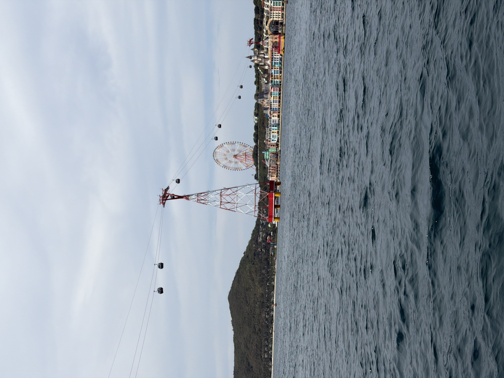 The image depicts a coastal scene, likely in or near Bac Nha Trang Ward, Vietnam. The right side of the frame is dominated by the ocean, showing a rippled surface. In the center of the frame stands a mountain with dense foliage. A large red and white cable car tower is prominently featured, with cable cars extending towards a distant Ferris wheel. The background also shows buildings in a vibrant color scheme, indicating a possible tourist area or resort. The sky is overcast with thin clouds.