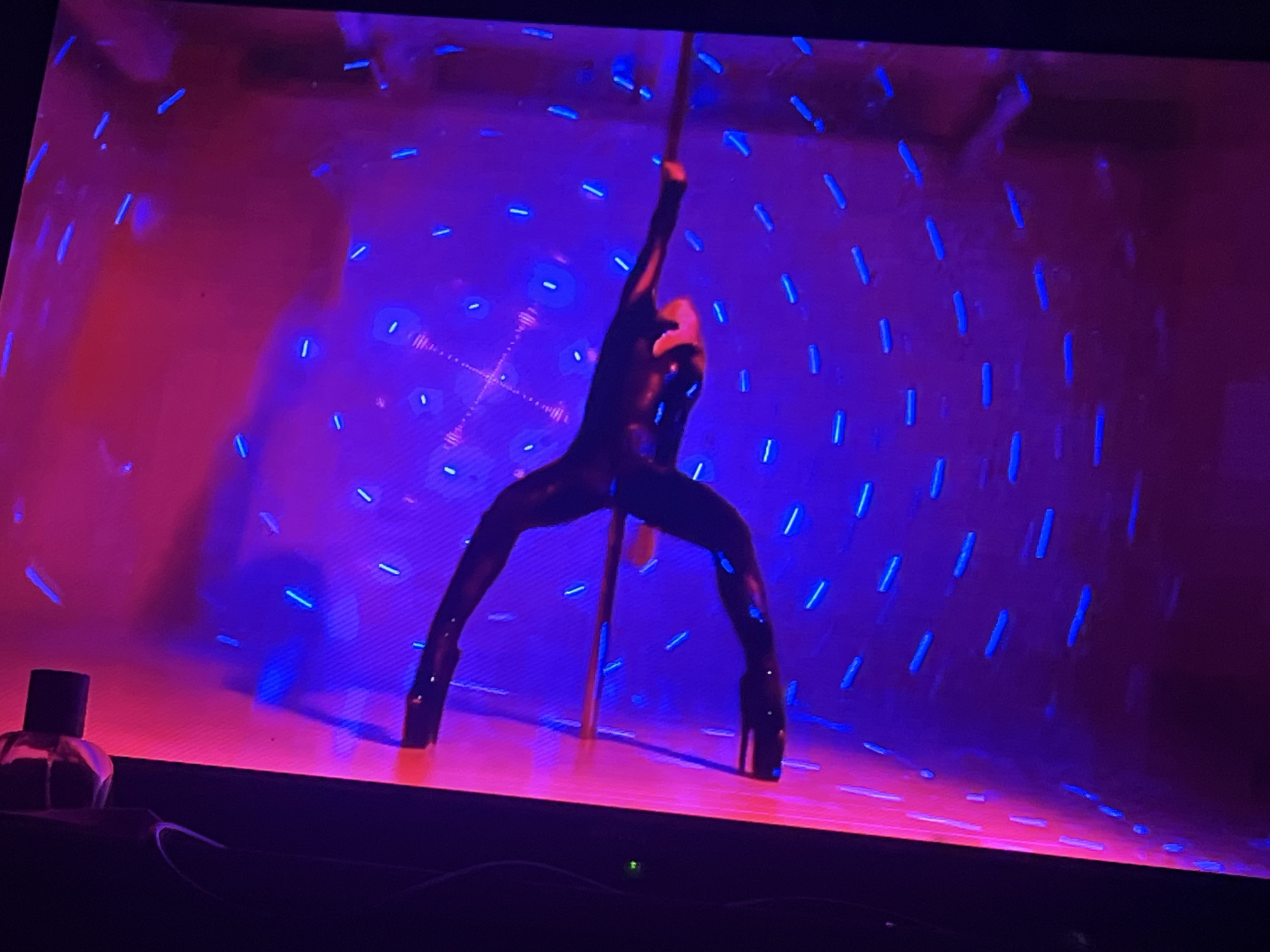 The image shows a performer in a black, form-fitting outfit on a pole, positioned in the center of a stage. The stage is illuminated with pink and purple lighting, with blue lights scattered across the background, creating a vibrant and energetic atmosphere. The performer's pose is dynamic, with legs spread wide and body arched. In the foreground, on the left side of the image, a bottle of perfume is visible on the edge of the stage. The scene appears to be a performance or entertainment venue. The image is not specific to Zoriane, Ukraine, as there are no discernible landmarks or context to associate it with that location.