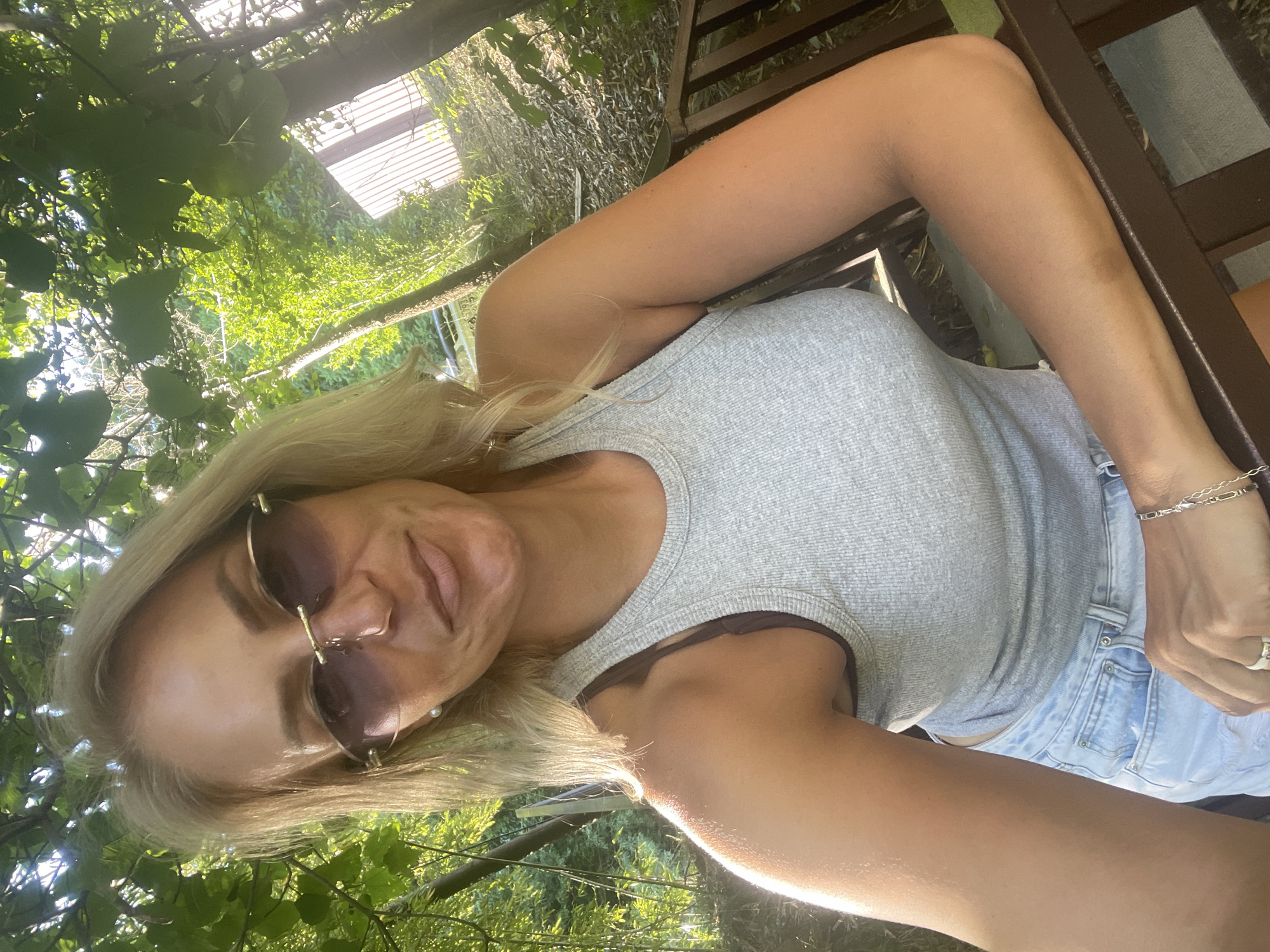 A blonde woman wearing sunglasses, a grey tank top, and denim shorts is sitting outdoors. She has a bracelet and a ring on her left hand. The background is green and lush with trees and foliage. The image was taken at a location in the city of Sochi, Russia.