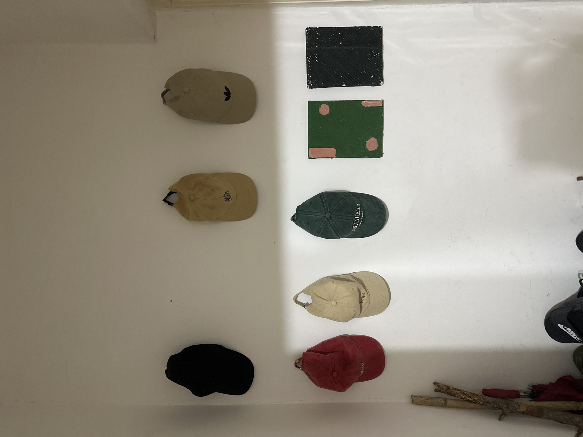 The image shows a white wall with various objects attached to it. On the left side, six baseball caps are aligned vertically. The caps are different colors: beige with an Adidas logo, tan, green with "Los Angeles" embroidery, and black. Adjacent to the caps, two square art pieces in black and green are on the wall. At the bottom right corner, parts of a motorcycle helmet and red material are visible. Below them, there is a small branch. The photograph seems to have been taken indoors. There is no indication of the location being in Budva, Montenegro.