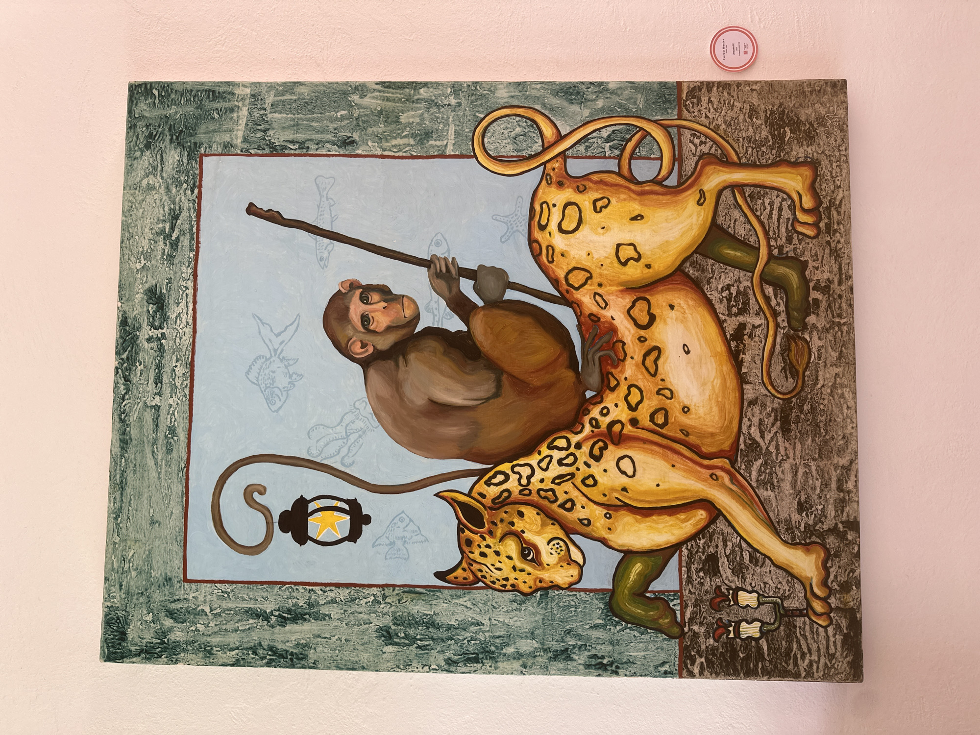 An illustrative painting, rich in texture and detail, hangs on a light pink wall. The artwork centrally features a monkey and a leopard-like creature within a framed composition.
The monkey, depicted in shades of brown with a lighter face and chest, is positioned on the left side of the canvas. It grips a dark brown stick with both hands, appearing to be suspended or swinging, and looks directly towards the viewer with an alert expression. The stick extends horizontally across the upper-middle section of the painting.
Intertwined with the monkey and the stick is a golden-yellow, spotted feline, resembling a leopard or jaguar. Its body wraps around, with its head facing downwards on the lower right and its tail extending upwards and curling. Some of its limbs are depicted in a greenish hue.
The background of the central scene is a pale blue, subtly adorned with faint, light-colored outlines of various marine life, including different types of fish, a starfish, and an octopus or squid. In the lower-left corner of this blue background, a dark brown, stylized lantern-like object with a bright yellow star design inside is visible.
The central painted image is enclosed by a textured border. The left and bottom sections of this border are rendered in a rough, greenish-blue pattern, suggestive of cracked ice or aged stone. The right and top sections feature a contrasting dark grey texture, possibly representing bark or rugged rock.
A small, circular red sticker is affixed to the wall above the painting's upper right corner. Visible text on the sticker includes "CYPREX ARTES", "MUSEUM...", and "ARANXA E". The painting's internal setting offers no specific cues for time of day or weather, presenting an abstract or ethereal environment.