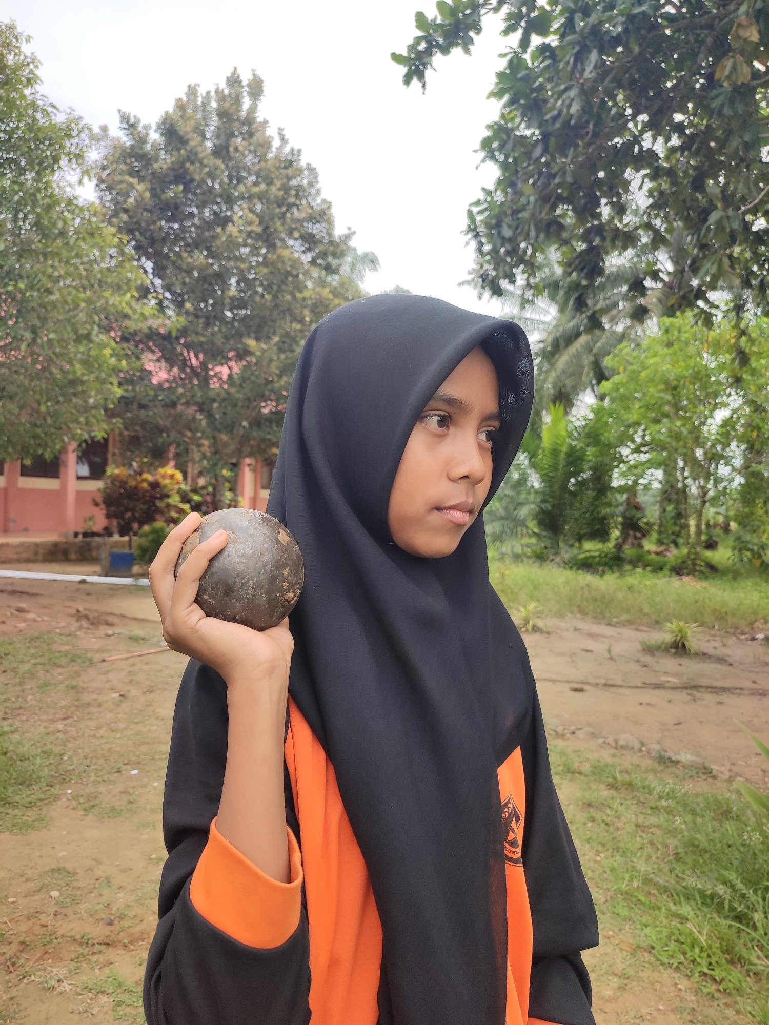 A young person wearing a black hijab and an orange shirt is holding a round, dark-colored object, possibly a stone or a ball, in their right hand. They are looking to their right, with a neutral expression. The person is standing outdoors in what appears to be a schoolyard or park. In the background, there is a building with a pinkish-orange facade, trees with green foliage, and a cloudy sky. The ground is covered with dirt and some patches of grass. The overall setting suggests a location in Blangriek, Indonesia, likely during daytime.
