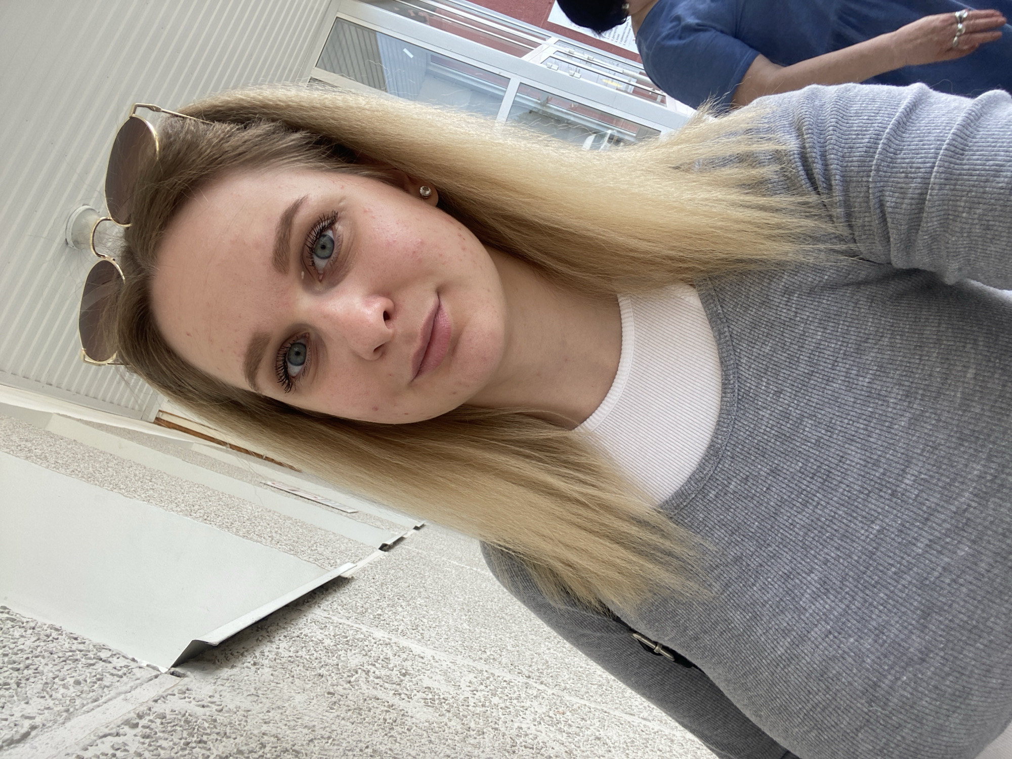 A young blonde woman wearing a grey long-sleeved shirt over a white undershirt, with sunglasses perched on her head, poses for a selfie. She has blue eyes and light skin with some redness. She is standing in an outdoor hallway with a textured, light-colored wall on the left and glass doors and a white ceiling overhead. Another person in a blue shirt is visible to the right, out of focus. The location is identified as Chaikovsky, Russia.