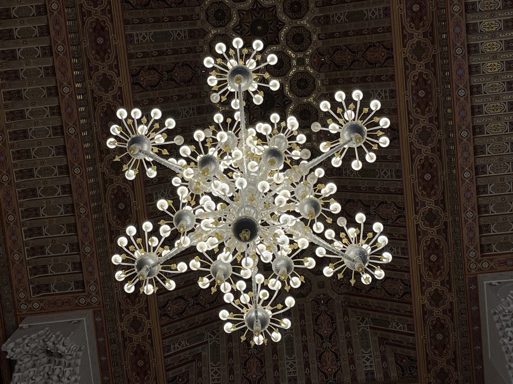 The image displays a large, ornate chandelier suspended from a richly decorated ceiling within an indoor architectural setting. The chandelier is the central subject, featuring a complex, multi-tiered design crafted from clear glass or crystal and metallic elements. Numerous arms radiate outwards from central spheres, each terminating in a small, illuminated, spherical bulb, creating a bright, starburst-like cluster of lights.
The ceiling provides a detailed backdrop, covered in intricate geometric patterns and stylized floral motifs. These patterns are rendered in a deep color palette dominated by dark reds, browns, golds, and hints of blue, indicative of traditional Moroccan or Islamic architectural decoration. The surface appears to be carved wood or meticulously painted plasterwork.
The bright illumination from the chandelier strongly contrasts with the darker, highly detailed ceiling, emphasizing both the fixture and the overhead artistry. The scene suggests the opulent interior of a grand building, such as a mosque, palace, or other significant edifice, aligning with the architectural traditions of Casablanca, Morocco. Some cropped sections reveal white, intricately carved plasterwork on a wall or architectural element in the corner, further underscoring the decorative nature of the environment. No people or visible text are present in the image.