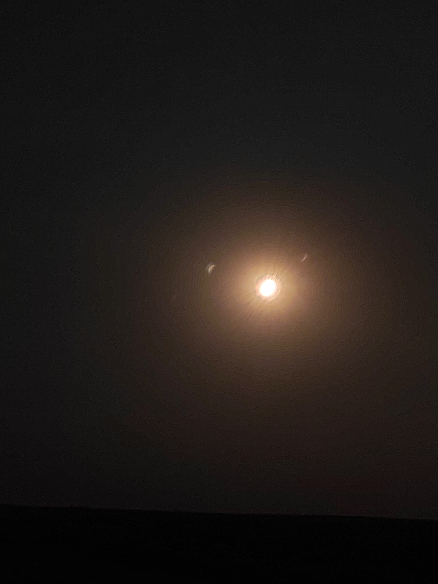 The image depicts a night sky with a bright, glowing celestial body, possibly the moon or sun, prominently featured in the center. The light source is surrounded by a soft halo or atmospheric effect, creating a diffused glow. To the left and right of this central light, there are two crescent shapes, also illuminated. The lower portion of the image is a dark, silhouetted landscape, suggesting a horizon line. The overall impression is one of a celestial event occurring over a dark terrain. The specific location in Autillo de Campos, Spain, cannot be determined from the visual information provided in the image.
