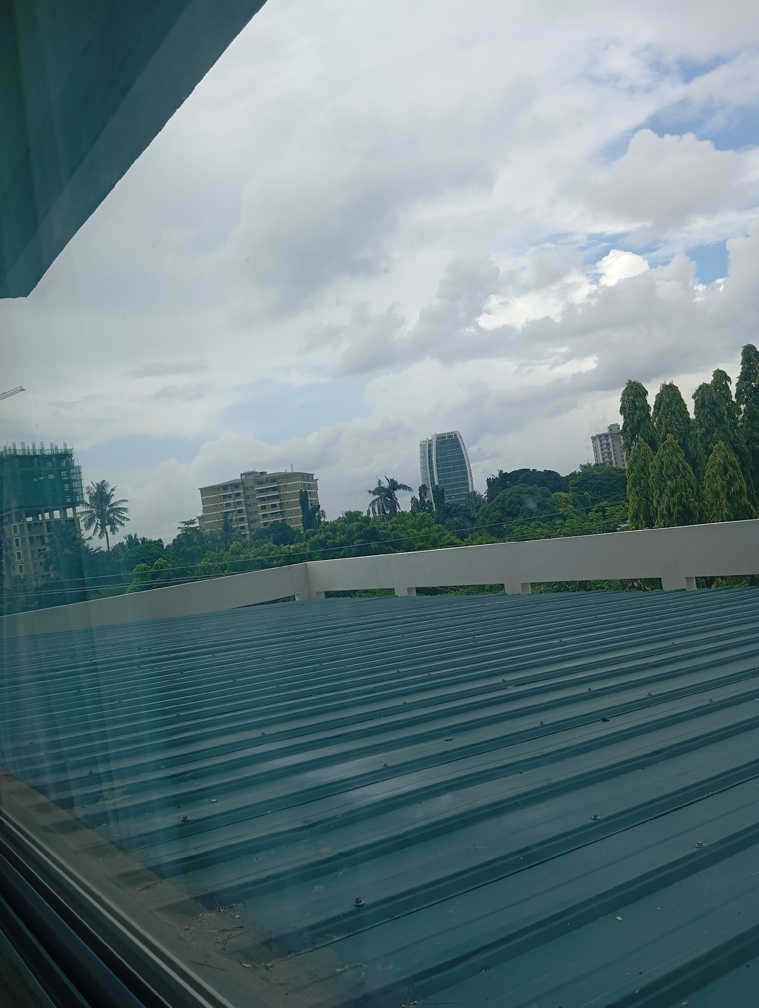 The image depicts an elevated view of a cityscape in Dar es-Salaam, Tanzania, observed through a window or from a vantage point overlooking a dark green, corrugated metal roof that occupies the lower half of the frame. The roof, textured with horizontal ridges and visible fasteners, shows minor surface wear.
Above the roof, a light-colored, possibly concrete or painted metal, parapet or railing extends horizontally across the midground. Beyond this barrier, a dense canopy of green trees and foliage forms a significant portion of the background.
The skyline is characterized by several multi-story buildings and structures under a partly cloudy sky, featuring white and grey cumulus clouds with visible patches of blue.
On the far left, a multi-story building under construction is visible, wrapped in green netting, with a tower crane extending above it. Adjacent to this construction, a tall palm tree stands. Towards the center-left, a completed, light-colored (beige or light grey) multi-story residential or commercial building is prominent, surrounded by abundant greenery. In the center-right, a distinctive, modern, white, curvilinear high-rise building, identifiable as the PSPF Towers due to its unique architectural design, rises above the tree line. To the far right, a cluster of tall, slender trees with dense green foliage partially obscures another high-rise structure. No individuals or specific actions are discernible in the scene. The overall impression is an urban environment undergoing development, characterized by a mix of modern architecture and substantial natural vegetation.