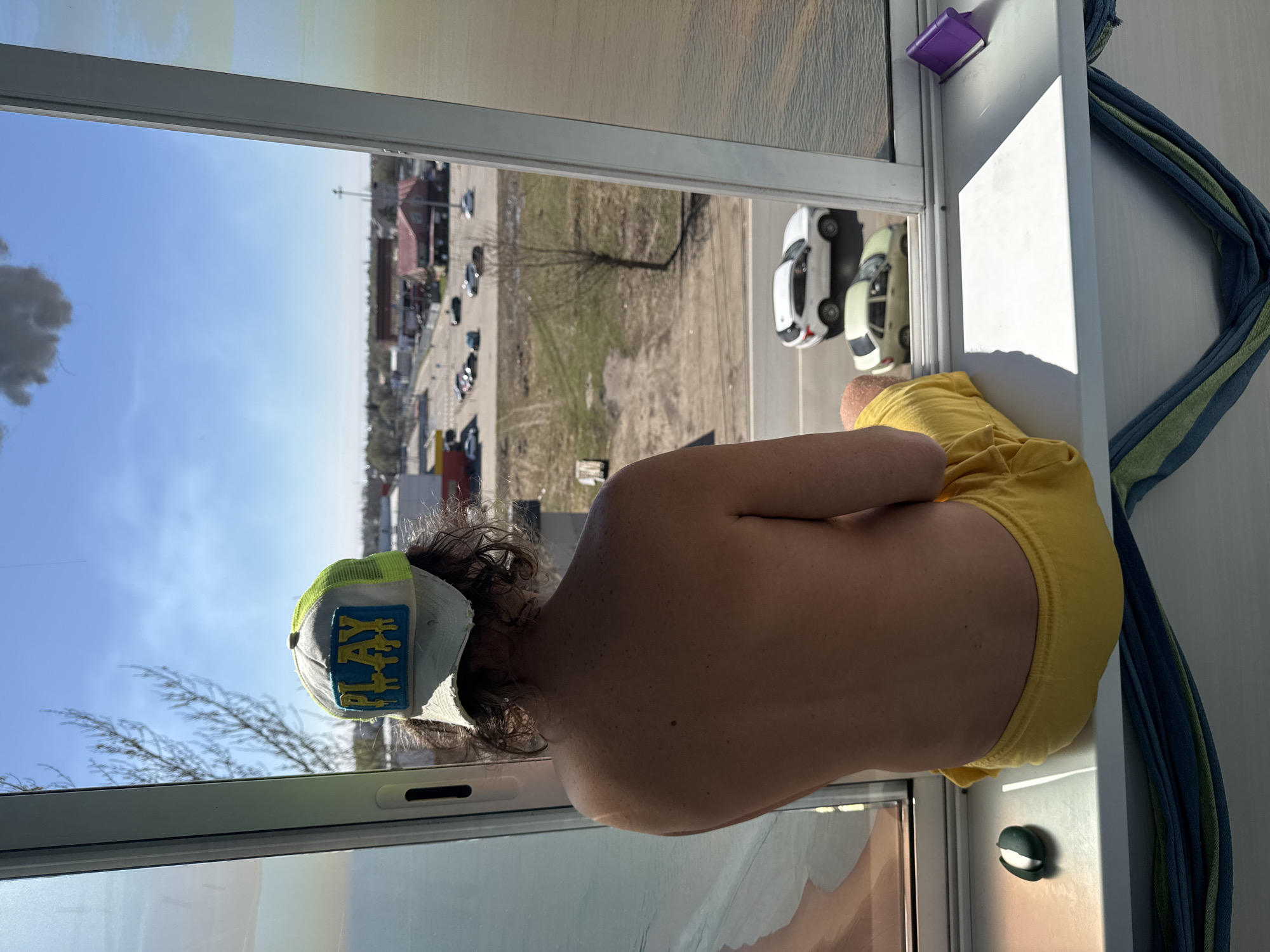 A person wearing a "PLAY" baseball cap and yellow underpants is sitting by a window, looking out over a scene featuring multiple cars, buildings, and open land. The location appears to be Novo-Nikol'skoe, Russia, as indicated by the image's metadata. A couple of toy cars sit on the windowsill.