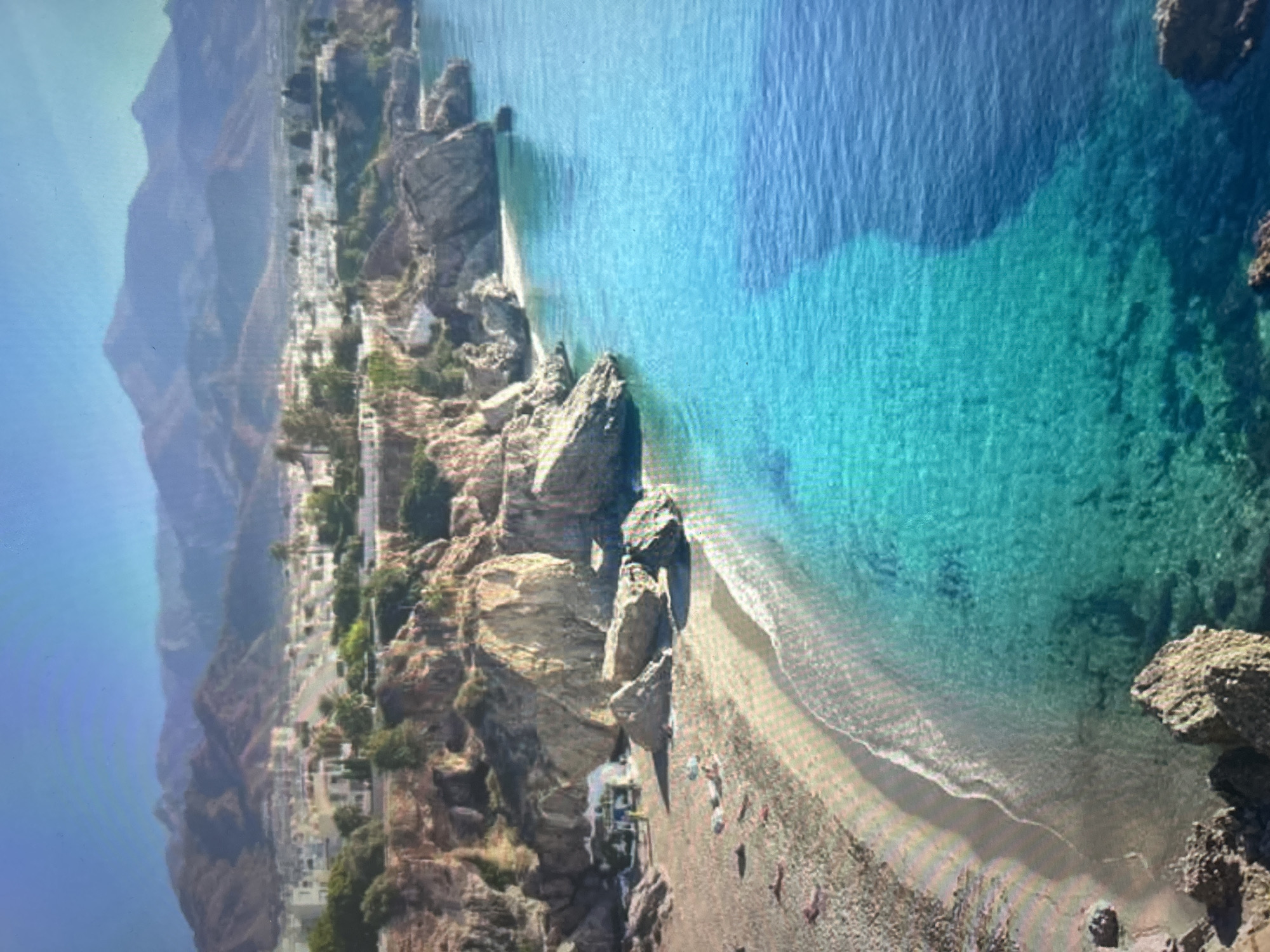 This aerial image depicts a vibrant coastal landscape, rotated 90 degrees clockwise from its natural orientation. The scene is dominated by rugged cliffs, a small town built into the hillside, and a stunning turquoise sea under a bright, clear sky.
On the left side (when correctly oriented), a series of towering, hazy mountains form a dramatic backdrop to the coastline. Below them, a town or village is integrated into the steep, rocky cliffs. Numerous light-colored buildings, possibly homes or hotels, are visible, many featuring red-tiled roofs, interspersed with patches of green vegetation and trees.
At the base of the cliffs, a sandy and pebbly beach curves along the shoreline. Small, indistinct figures, presumably people, are scattered on the beach, some near colorful beach umbrellas, suggesting leisure activities like sunbathing or relaxing by the sea. A small structure, possibly a beach hut or cafe, is discernible near the waterline amidst a cluster of large rocks.
The right side of the image features the calm, crystal-clear waters of the sea. The water exhibits a beautiful gradient, shifting from a bright, shallow turquoise near the shore, where the sandy and rocky seabed is visible, to a deeper, darker blue further out. Small waves can be seen gently breaking on the beach. The overall impression is one of a serene and picturesque coastal environment, basking in bright daylight. No discernible text is visible.