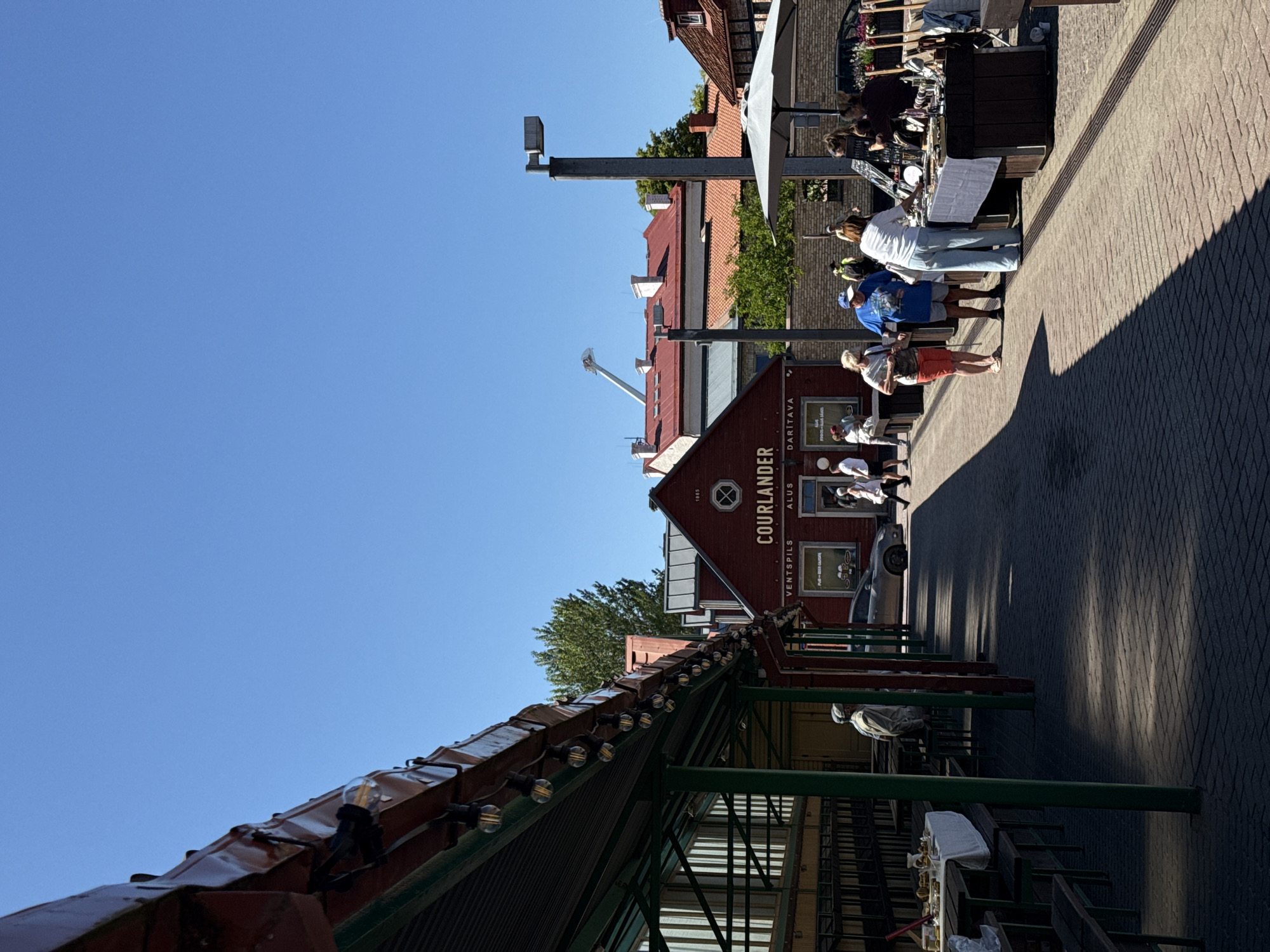 This is a ground-level, medium-shot, outdoor photograph taken on a sunny day in Ventspils, Latvia. The image captures a street scene with a covered market area on the left and market stalls on the right. The ground is paved with light-colored brickwork.
On the left, a long, covered structure with a dark red roof and green pillars extends into the foreground. This structure appears to be an open-air market or seating area, with tables and benches set up inside. A person in a light-colored jacket and cap is standing in this area, looking away from the camera.
In the middle ground, a prominent red building with white trim bears the signage "1965 COURLLANDER" and "VENTSPILS ALUS DARITAVA". A car is parked in front of this building. Several people are walking or standing near the building.
On the right side of the image, market stalls are set up. One stall, covered by a large white umbrella, displays various items, possibly jewelry or crafts, on a dark blue tablecloth. Several people are gathered around this stall, with one woman appearing to examine the items, while a vendor is interacting with customers.
The sky is a clear, bright blue. The background consists of other buildings with red and white roofs, and trees. The overall atmosphere suggests a vibrant market or a town center on a warm day.