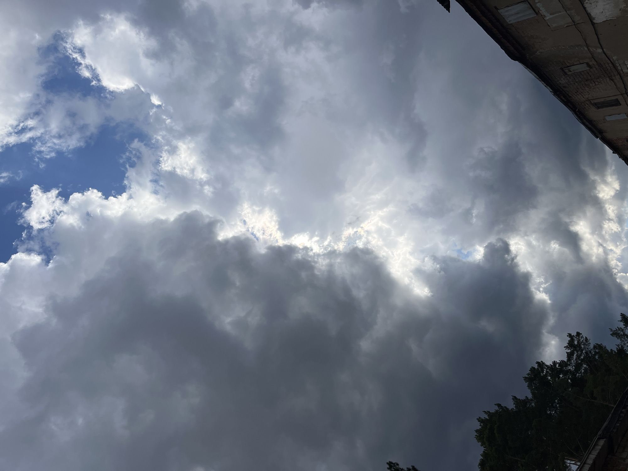 The image presents a dramatic view of a dynamic sky over Chaikovsky, Russia, dominated by an extensive cloudscape. The sky is filled with a mixture of clouds, ranging from bright, sunlit white formations to heavy, dark grey storm clouds. In the upper left quadrant, patches of clear, deep blue sky are visible, interspersed with wispy white clouds. The central and lower portions of the frame are largely obscured by thick, dark clouds, which appear dense and heavy, hinting at impending rain or an active weather system. Intense sunlight breaks through gaps in these darker clouds, creating brilliant white highlights and a sense of light pushing through gloom.
Towards the bottom right, the dark, dense foliage of trees is visible, silhouetted against the clouds, suggesting an outdoor, possibly urban or suburban setting. In the upper right corner, a segment of an old, weathered building structure is visible. It features a visible roofline or ledge, and what appear to be obscured or bricked-up window openings, contributing to the architectural context. The building's aged appearance suggests a long-standing structure.
The scene captures a moment of active weather, with the interplay of bright sunlight and dark clouds creating a visually striking and dynamic atmosphere. The time of day is likely mid-day to late afternoon, given the intensity and direction of the light breaking through the clouds. There is no visible text in the image.