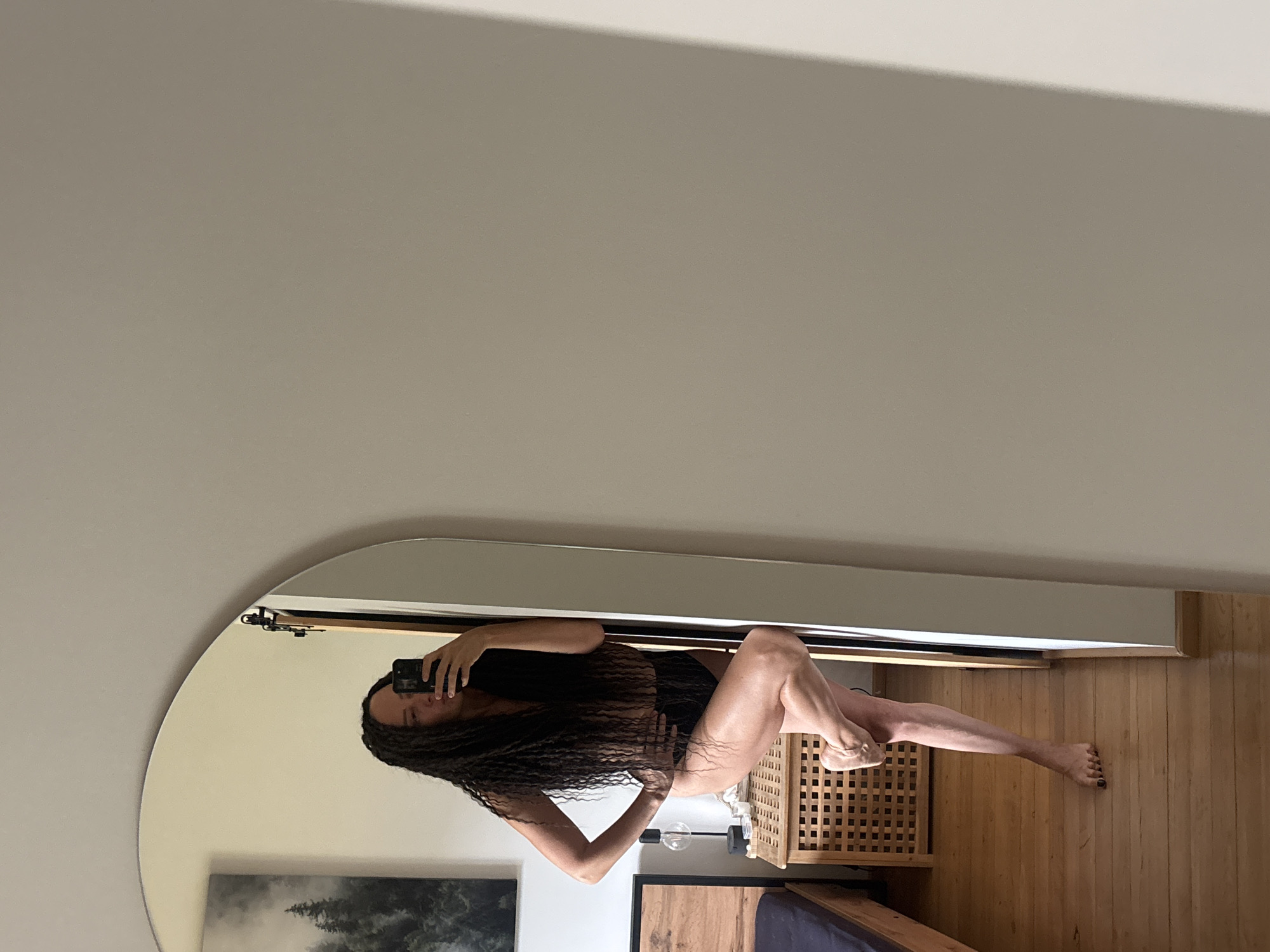 A woman is taking a selfie in a mirror. She is wearing a dark-colored garment. The room contains a wooden floor, a small cabinet, a bed, a painting on the wall, and a light fixture. There is no evidence to support the claim that this image was taken in Chaikovsky, Russia.